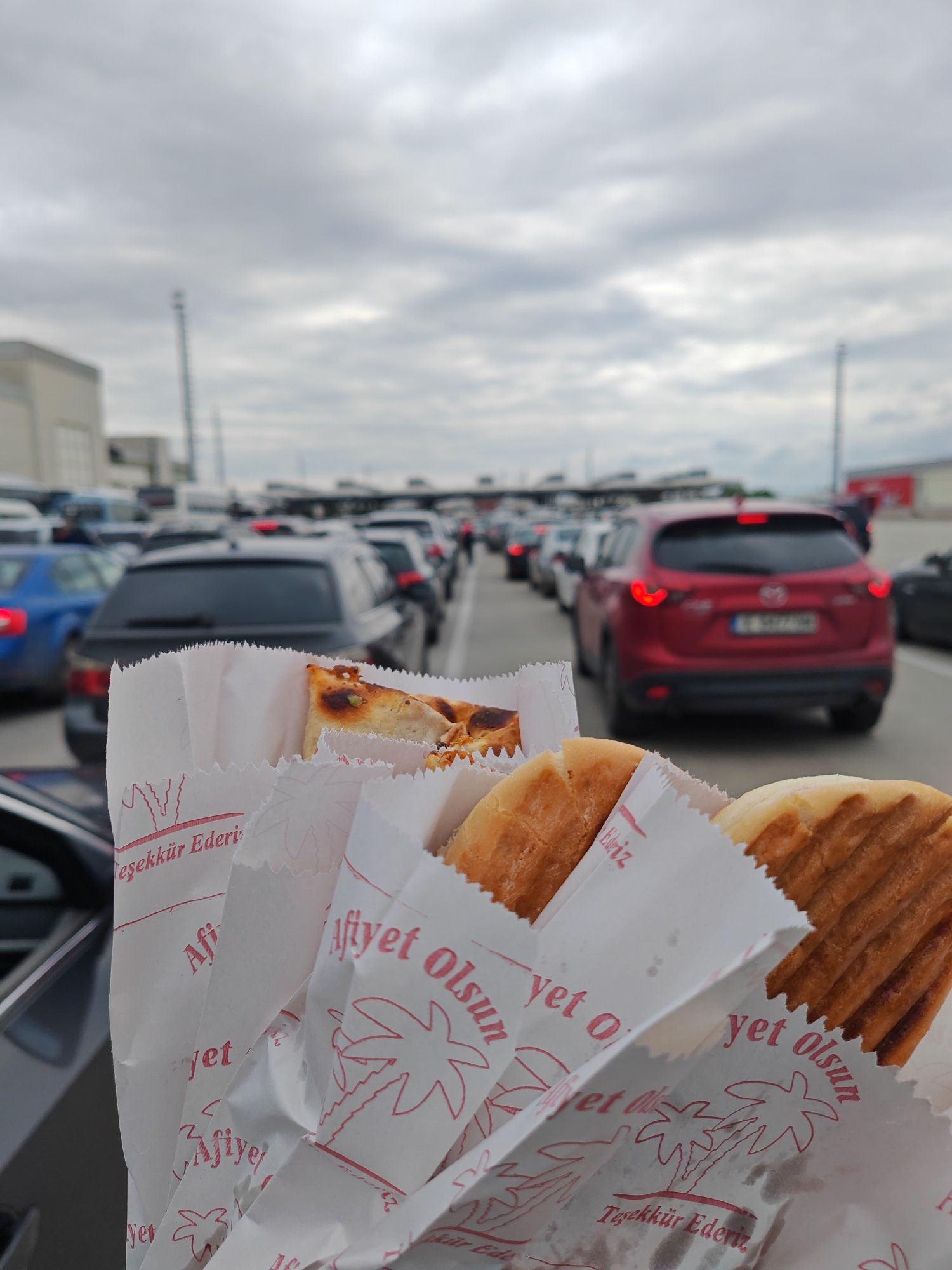 The photograph captures a first-person perspective from inside a vehicle, focusing on several toasted sandwiches held in white paper wrappers in the foreground. The wrappers are adorned with a red design featuring palm trees and Turkish text, including "Afiyet Olsun" (Enjoy your meal) and "Teşekkür Ederiz" (Thank you). One wrapper also partially shows "Edriz."
In the background, a long queue of cars stretches into the distance under an overcast sky. The cars are stationary, with brake lights visible on many, suggesting a significant wait. Notable vehicles in the line include a blue sedan on the left, a dark grey sedan in the center, and a prominent red Mazda CX-5 SUV on the right. The setting appears to be a multi-lane border crossing or checkpoint, given the context of Kapitan Andreevo, Bulgaria, a major crossing point. The cloudy weather indicates a daytime scene. The act of eating the sandwiches implies that the occupants of the vehicle are passing the time during a prolonged wait, a common occurrence at busy border points.
