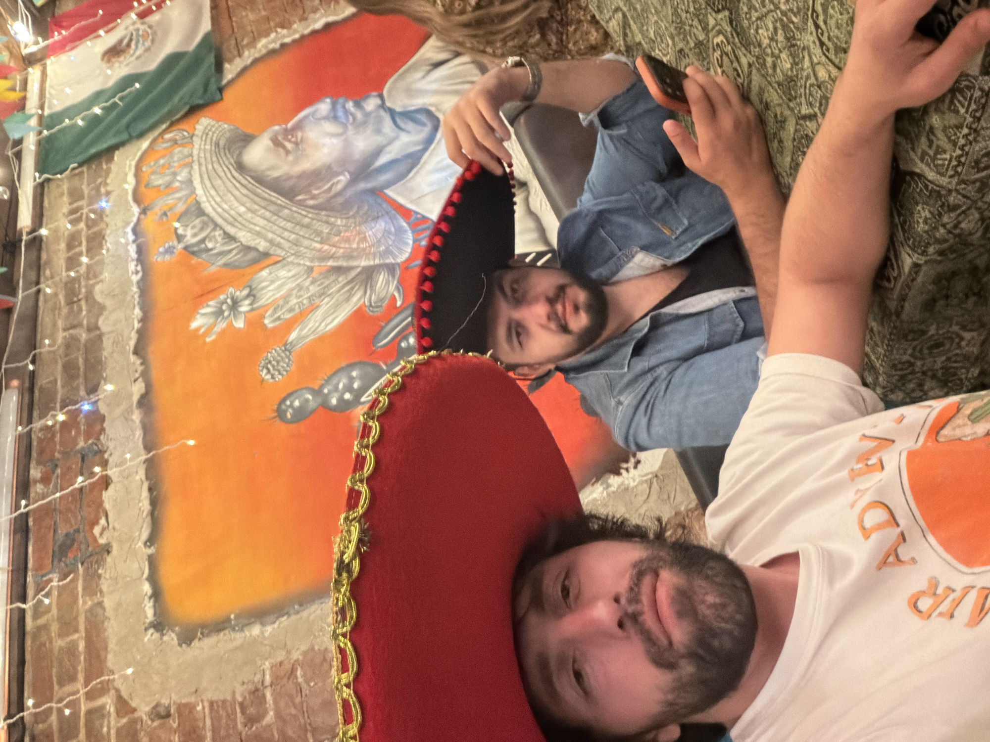 Two men are lying down next to each other. One man wears a red sombrero and a white shirt with an orange graphic. The other man wears a black sombrero with red trim and a blue denim jacket. They are positioned in front of a mural depicting a man in a sombrero, next to a Mexican flag. The mural is painted on a brick wall adorned with string lights. The location is likely a restaurant or bar with a Mexican theme, possibly in Krasnoyarsk, Russia.