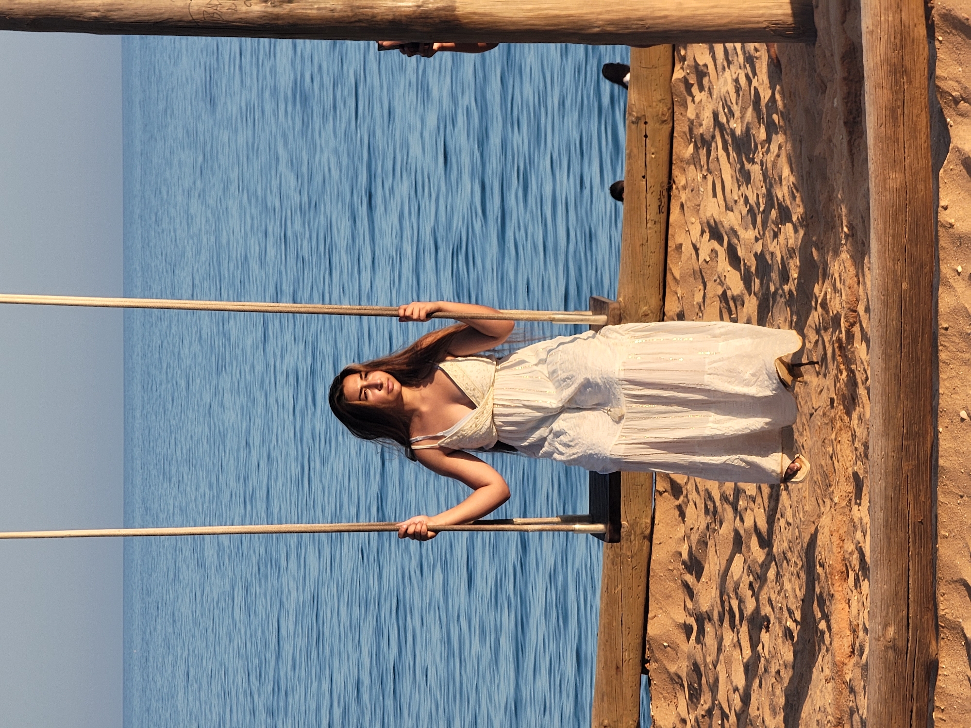 A young woman with long, dark hair is sitting on a wooden swing by the sea in Quarteira, Portugal. She is wearing a long, white, sleeveless dress with a V-neck and spaghetti straps. The dress has a tiered skirt and is adorned with subtle gold detailing. Her feet, clad in open-toed sandals, rest on the sandy ground. The swing is suspended by two thick ropes, and the wooden seat is visible beneath her.
The background is dominated by the calm, blue sea, which stretches to the horizon under a clear, pale blue sky. The shoreline is made up of light brown sand, with scattered pebbles and shells. In the foreground, rough wooden planks form the structure of the swing's platform. The scene is illuminated by the warm, golden light of what appears to be late afternoon or early morning sun, casting long shadows across the sand. The overall impression is one of peaceful solitude and natural beauty.