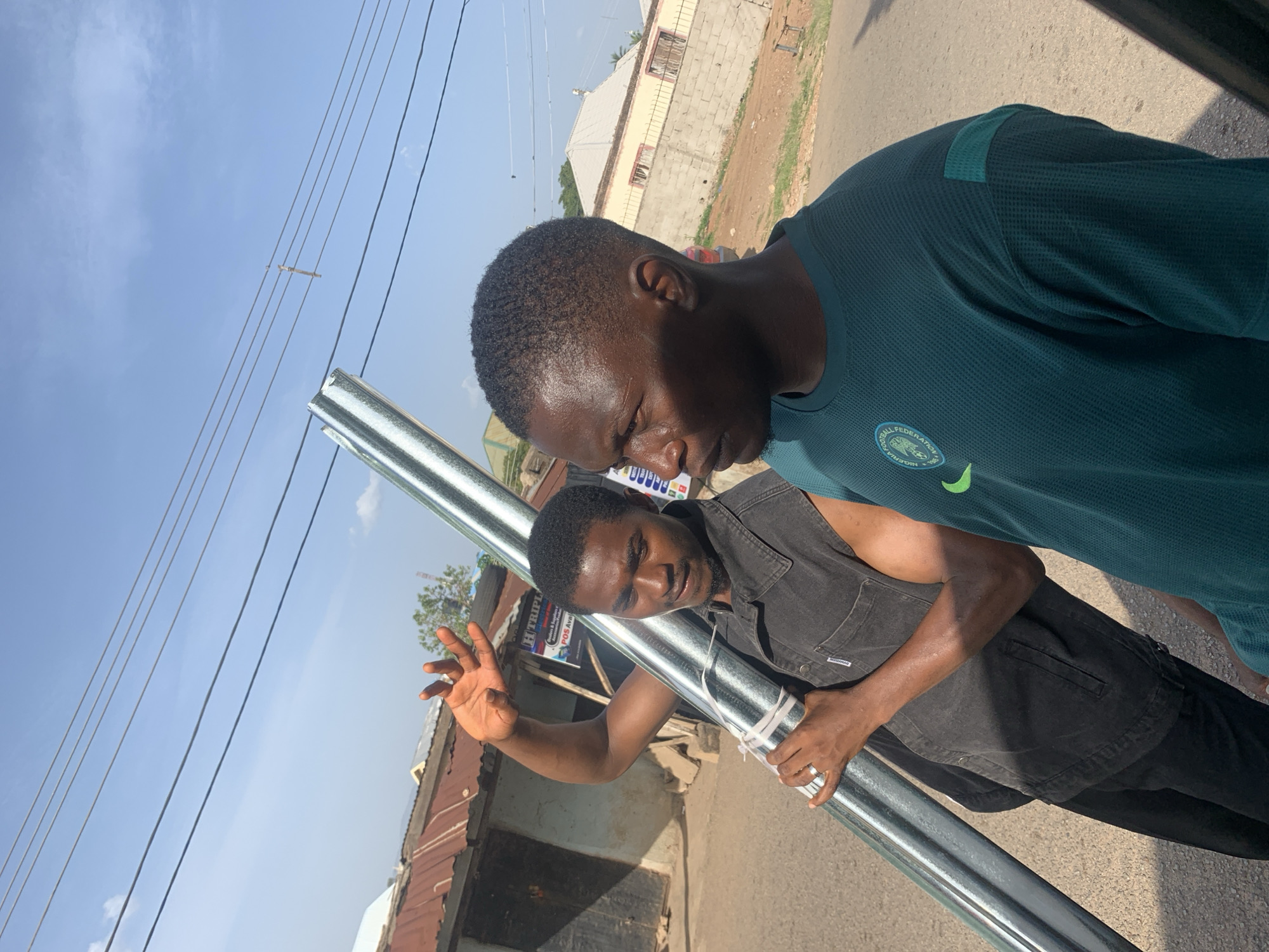 Two young men are carrying a long, cylindrical metal pipe outdoors, likely on a street in a suburban area of Jos, Nigeria. The man closer to the camera wears a dark gray vest, while the other wears a green sports shirt with a logo indicating the Nigeria Football Federation. The man in the vest holds the pipe with one hand while extending the other, palm outward, as if directing someone. The environment includes buildings with corrugated metal roofs and overhead electrical wires, typical of the Jos urban landscape. A sign for "H Triple" is visible in the background, indicating a local business. The street surface is visible. The overall scene suggests they are transporting or installing the pipe.
