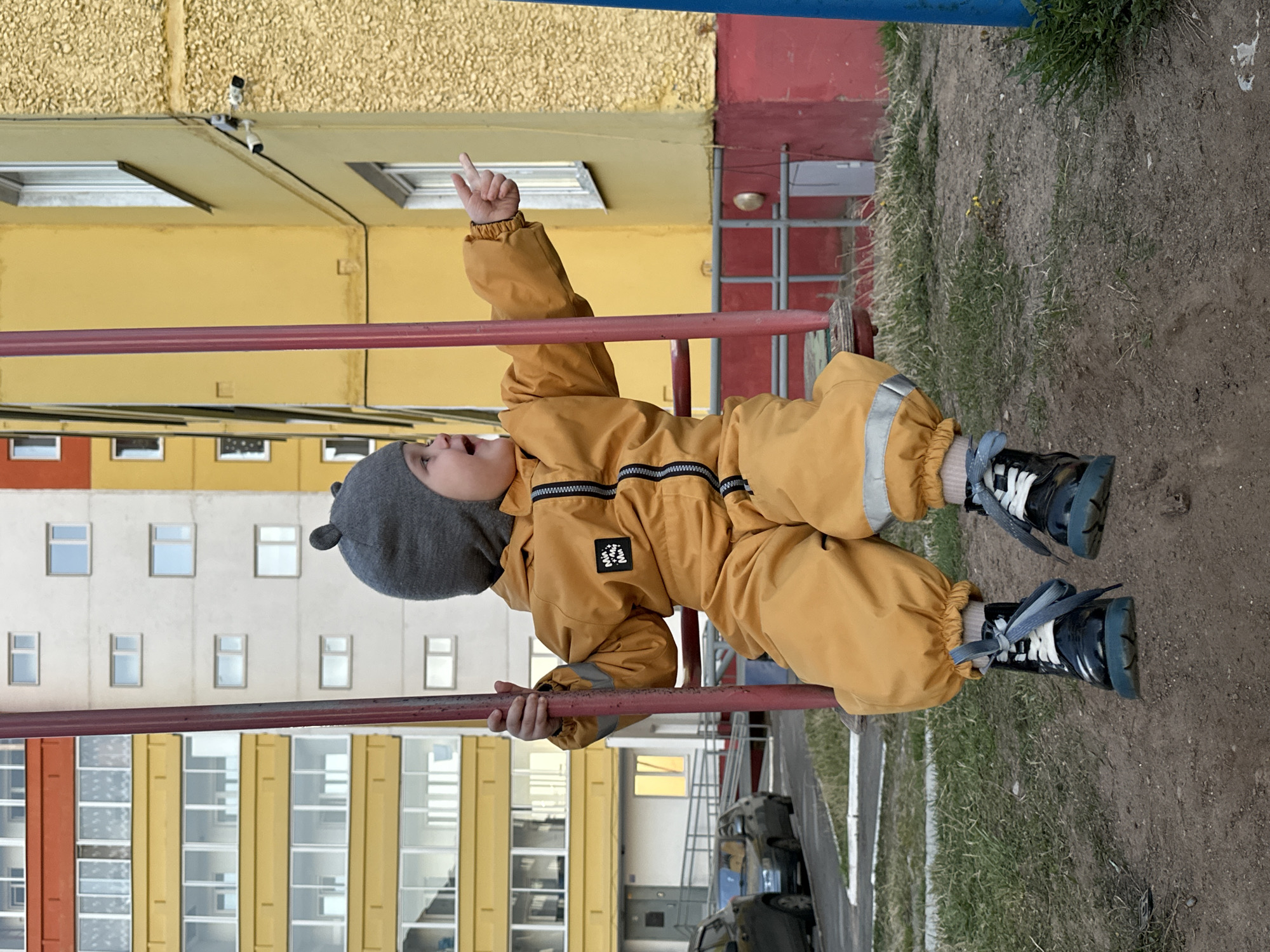A young child, likely a toddler, wearing a yellow jumpsuit and a gray hat with ear-like protrusions, is suspended on a red metal bar, possibly part of a playground structure, outdoors. The child is holding onto the bar with both hands and has one arm extended, index finger pointing upwards. The child is wearing black and white shoes.
The backdrop reveals a multi-story building with a mixed facade: sections are painted yellow, white, red, and a blue vertical element is present at the top right. Numerous windows are visible on the building. The ground beneath the bar and in the foreground is a mixture of soil and sparse grass. A partially visible vehicle is present in the lower right.
The location is likely in a residential area within the city of Chaikovsky, Russia, suggested by the architectural style of the building which is common in the region.