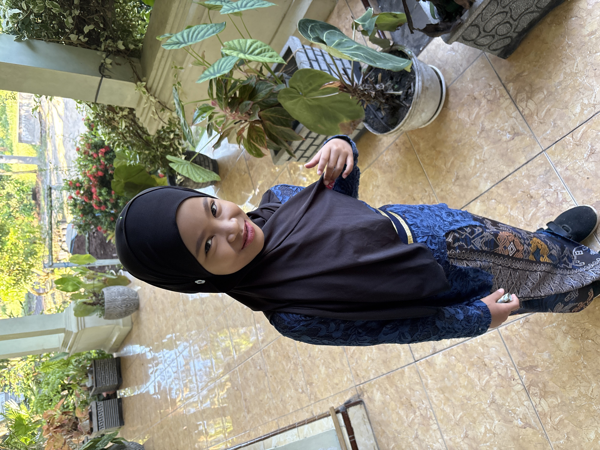 A young girl wearing a black hijab, a blue long-sleeved dress with lace details, and a patterned skirt is standing on a tiled floor. She has a slight smile on her face and appears to be looking directly at the camera. The environment includes various potted plants, some with large green leaves, located along what appears to be a porch or veranda. The setting suggests an outdoor or semi-outdoor space. The location is in Gadingrejo, Indonesia. The girl is holding what appears to be a small, rolled-up object in her right hand.