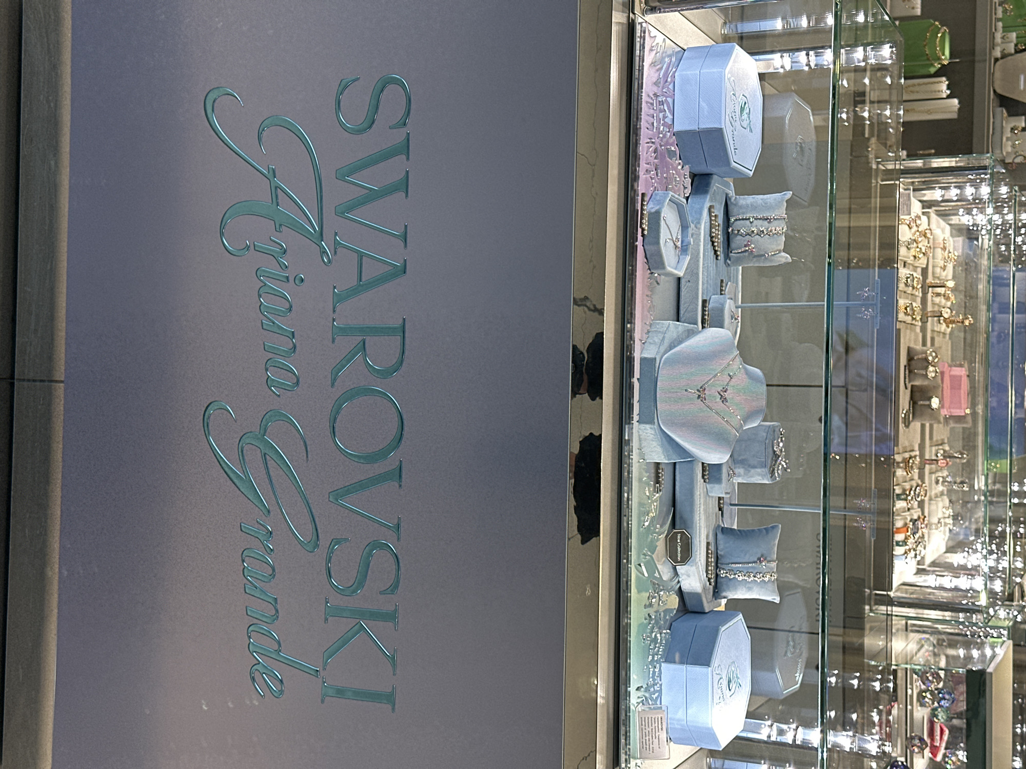 The image shows a jewelry display case with "Swarovski Ariana Grande" written vertically in a blue-green serif font on the left side of the image. The right side shows a glass jewelry case filled with various pieces of jewelry in blue display boxes and on blue stands. The lighting is bright, and reflections can be seen on the glass surfaces. A deeper view into the display case shows further rows of jewelry, some with gold and pink colors visible. The location, Sofia, Bulgaria, is not discernable from the image.