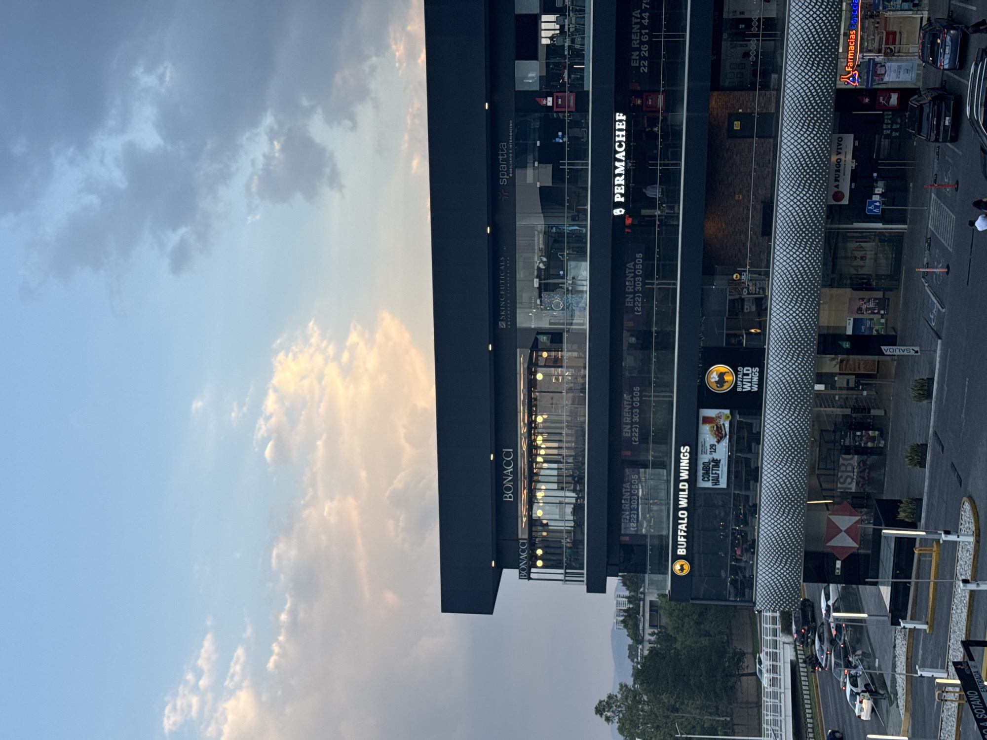The image depicts a modern commercial building in San Bernardino Tlaxcalancingo, Mexico, under a partly cloudy sky during what appears to be the late afternoon or early evening. The sky transitions from a soft blue to warmer tones, with some clouds illuminated by the setting or rising sun, while artificial lights are already on inside the building.
The multi-story structure features large glass windows and dark-colored facades, housing various businesses. Visible signs identify tenants such as "BONACCI," "SKINCEUTICALS ADVANCED CLINICAL SPA," "spartta MOBILIARIO E INTERIORISMO," "PERMACHEF," and "BUFFALO WILD WINGS," which advertises a "COMBO HALFTIME 129." The logos for Buffalo Wild Wings and HSBC are also present. Several "EN RENTA" (For Rent) signs with phone numbers, including "(222) 303 0505" and "22 26 61 44 79," indicate available commercial spaces.
A paved road runs in front of the building, with several cars parked or driving, suggesting typical daily activity. Road markings and signs like "ENTRADA" (Entrance), "SALIDA" (Exit), and "SOTANO" (Basement) are visible, indicating access points. Further down the street, "Farmacias Espe" and "A FUEGO VIVO" are identifiable businesses. In the distance, mountains or hills rise, providing a natural backdrop to the developed area. The overall scene is calm, characteristic of a commercial area at dusk or dawn.