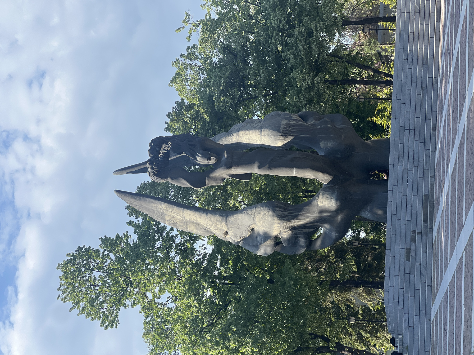 A towering, dark-colored monumental sculpture stands prominently in what appears to be a public square or park, set against a backdrop of green trees and a partly cloudy sky. The sculpture depicts a robust, stylized human figure, likely a soldier, with arms raised. One arm, possibly holding a weapon or another object, extends upwards. The material appears to be bronze or a similar weathered metal, showing subtle variations in light and shadow across its surface.
Surrounding the sculpture, tall trees with dense green foliage provide a natural setting. To the right of the sculpture, a section of a building or a large wall is visible, constructed from light grey rectangular blocks with distinct white grout lines, forming a grid pattern. The ground in the foreground and around the base of the monument is paved. The scene is brightly lit by natural daylight, indicating a clear or partly cloudy day. The overall impression is one of solemnity or commemoration. Based on the provided context, this is likely the "Monument to the Soviet Army" (often referred to as Alyosha) located in Plovdiv, Bulgaria. No text is visible in the image.
