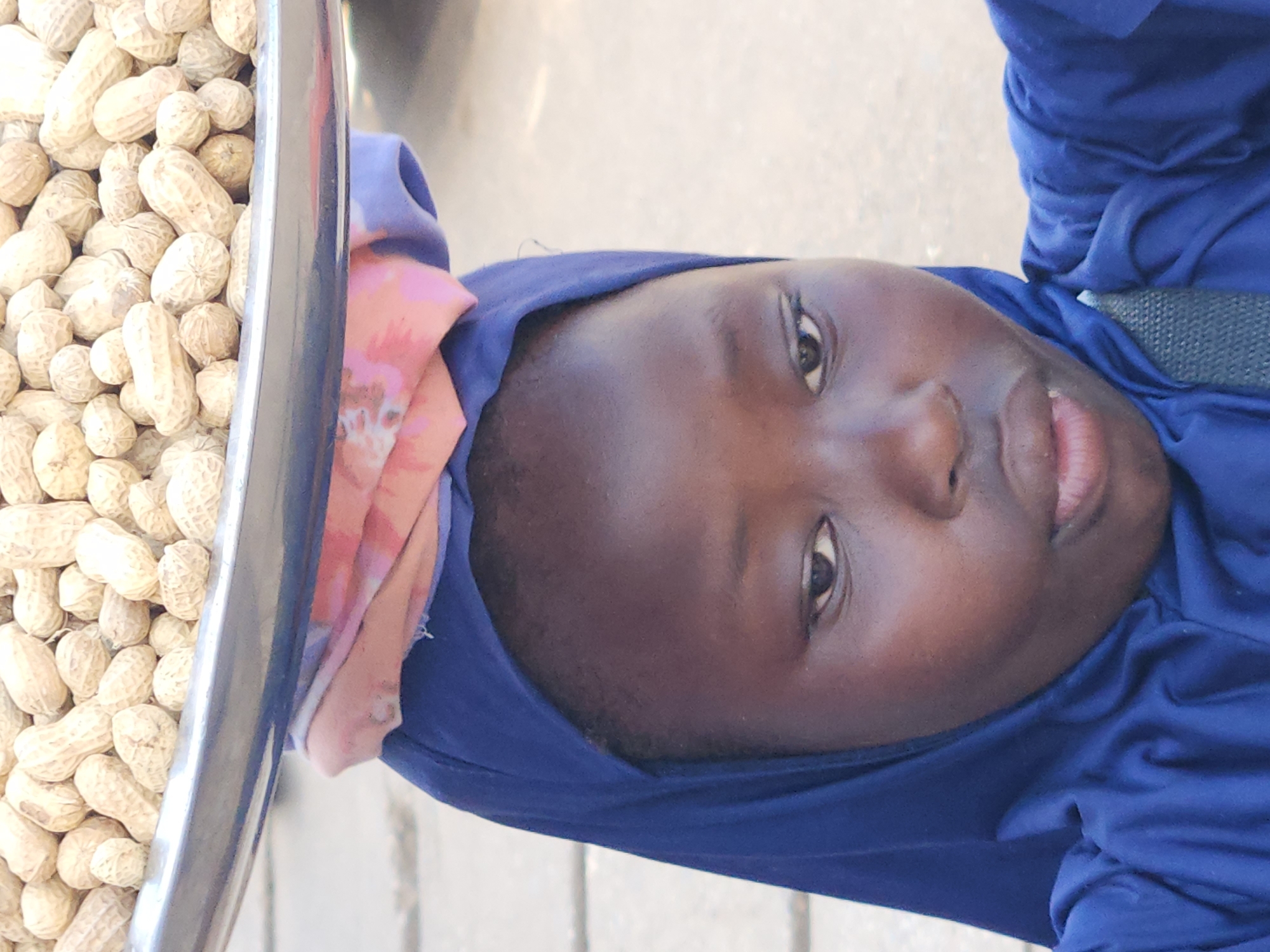 A young Black person is shown from the shoulders up, looking slightly to their left. They are wearing a blue headscarf that covers their hair and neck, and a matching blue garment. A colorful headwrap with pink, orange, and purple patterns peeks out from under the headscarf. They are balancing a large metal tray filled with peanuts on their head. The peanuts are in their shells and have a light brown, textured appearance. The background is blurred and appears to be an outdoor setting, possibly a street. The lighting is natural and appears to be daytime.
