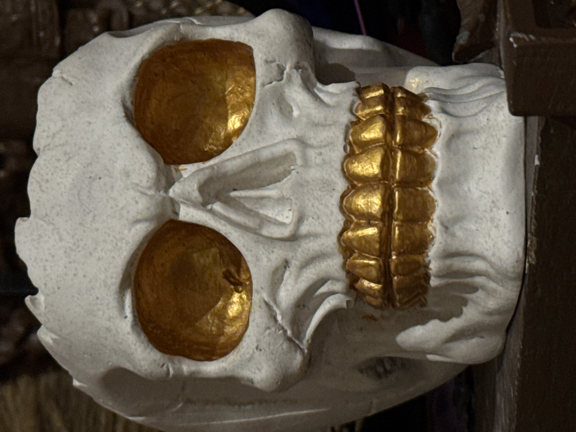The media file presents a close-up, vertically oriented image of a sculptural element. The primary subject is an abstract, skull-like or head-shaped object, rendered in a light-colored material, possibly plaster or aged stone, which exhibits a rough, porous texture with visible irregularities.
Two prominent, oval-shaped indentations, positioned where eyes would be, are filled with a shimmering, golden substance that reflects light. To the right of the central axis, a distinct rectangular golden element is present, composed of multiple segmented, convex sections, resembling a ribbed band or a stylized lower jaw.
The sculpture is set against a dark, out-of-focus background, which highlights the object. The lighting appears to be artificial, emphasizing the textures of the light material and the metallic sheen of the golden details. No activity is taking place, and no people or text are visible. The object serves as a decorative or symbolic piece. Given the location context of Novo-Nikol'skoe, Russia, it could be an architectural detail, an artifact, or part of an art installation.