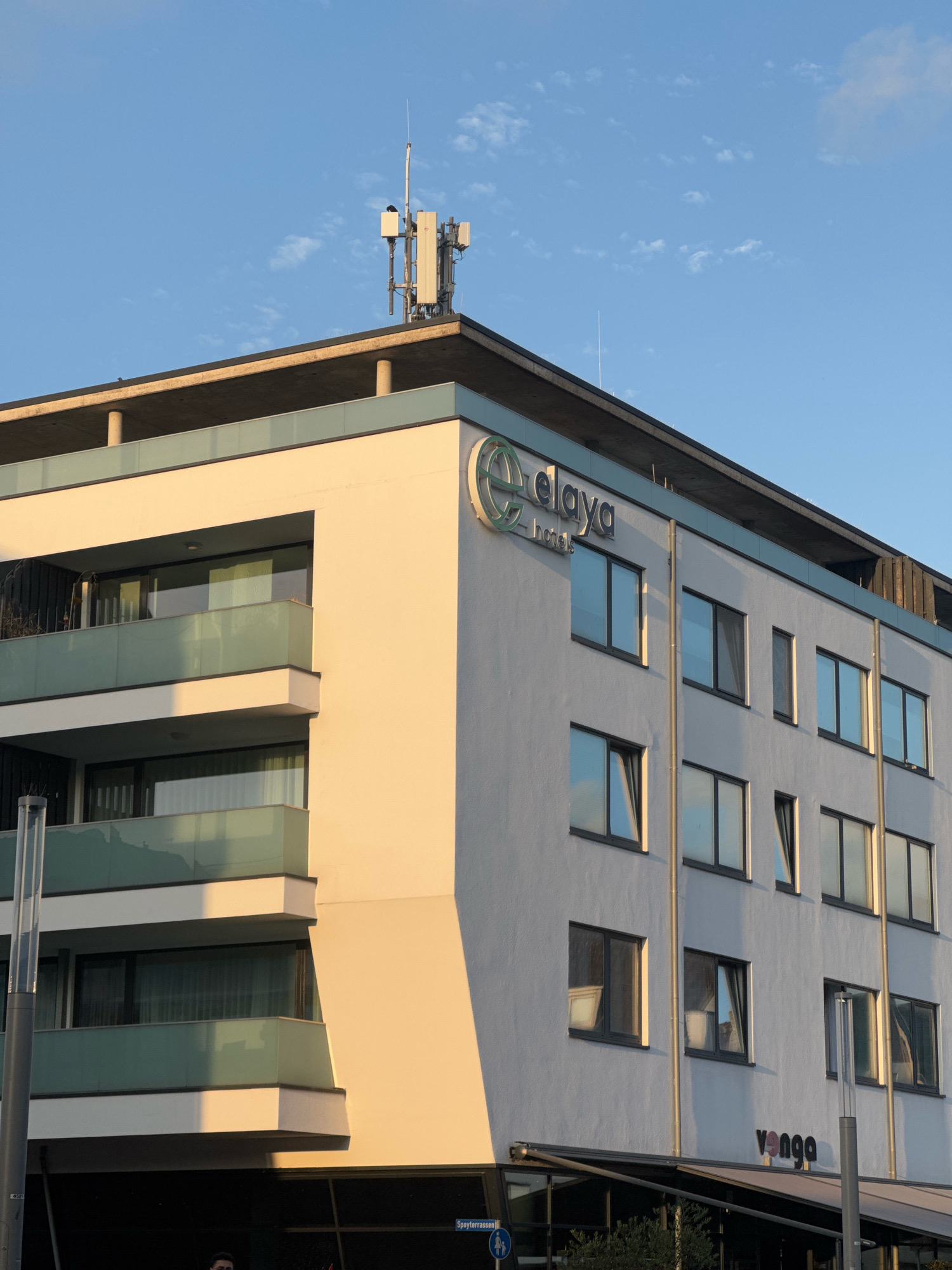 The image shows the "elaya hotels" building in Cleves, Germany. The building is modern with a white facade and features multiple balconies with glass railings. The "elaya hotels" logo is prominently displayed on the upper left side of the building. Below the hotel is a sign for "venga". A communication tower is visible on the rooftop. The sky is blue with scattered clouds, and the sun is casting warm light on the building. A street sign reading "Spyterrrasse" is visible in the lower right corner. No people are clearly identifiable in the image.