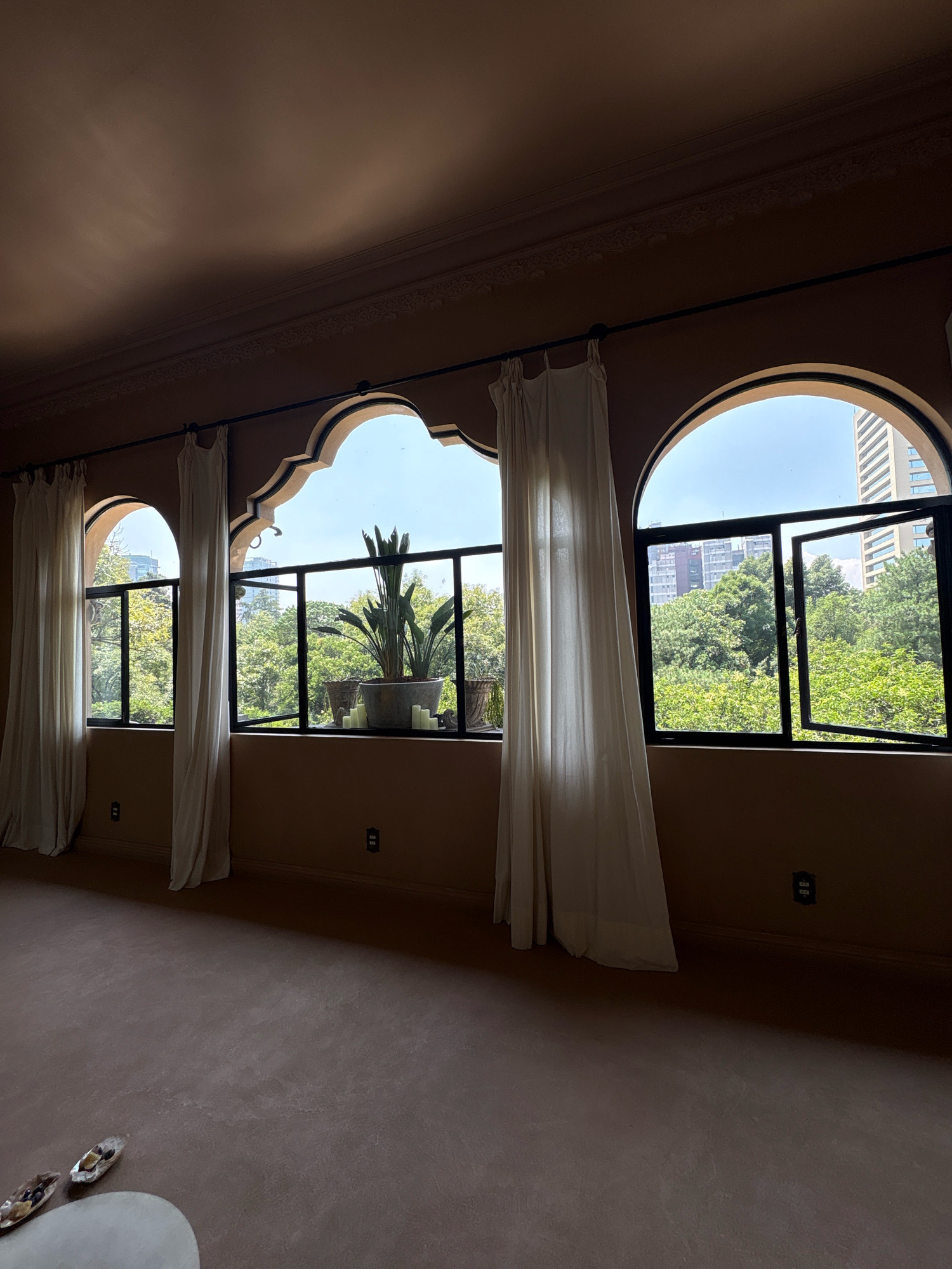 The image depicts an interior view of a room with distinctive arched windows offering a view of lush greenery and urban buildings in Mexico City, Mexico. The room has taupe-colored walls and a matching carpeted floor. Three arched windows are prominent, each with white curtains drawn to the sides. The central window is larger and features an ornamental plant in a pot, accompanied by several pillar candles. Through the windows, a dense canopy of trees is visible, interspersed with modern high-rise buildings. The sunlight streams in, casting shadows on the floor. On the floor in the foreground, two decorative objects resembling bowls are present, one containing dark berries. Two electrical outlets are visible on the lower part of the walls. The architecture of the windows suggests a blend of traditional and modern design elements.