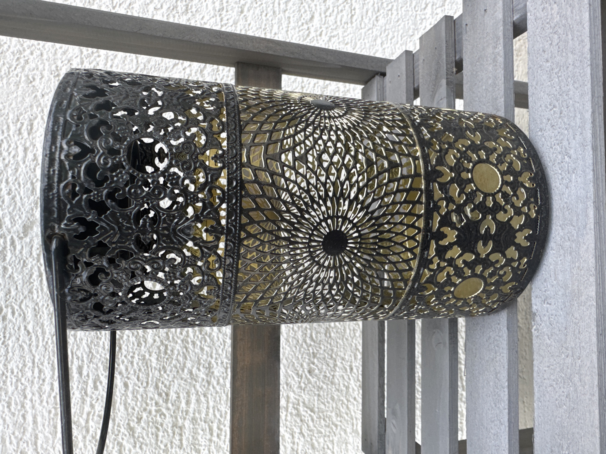 A cylindrical metal lantern with an intricate, filigree-like design hangs horizontally from a wooden structure against a textured white wall. The lantern is primarily dark gray or black with a metallic gold interior, visible through the cutouts. The design features three distinct sections: one with ornate scrolling patterns, a central section with a radiating sunburst motif composed of diamond-shaped openings, and a rightmost section with a repeating floral or organic pattern. The lantern appears to be a decorative outdoor item, possibly a solar-powered lamp, as evidenced by a black power cord that dangles from the left side and trails down towards the bottom of the frame. The wooden structure supporting the lantern consists of horizontal and vertical beams, painted in a weathered gray tone, suggesting an outdoor setting like a patio, balcony, or garden. The white wall behind is rough and textured, possibly stucco or concrete. The lighting suggests daytime, with soft shadows indicating ambient light rather than direct sunlight. No people are visible in the image. The overall impression is one of decorative illumination and outdoor ambiance. The location context is Hohenbrunn, Germany.