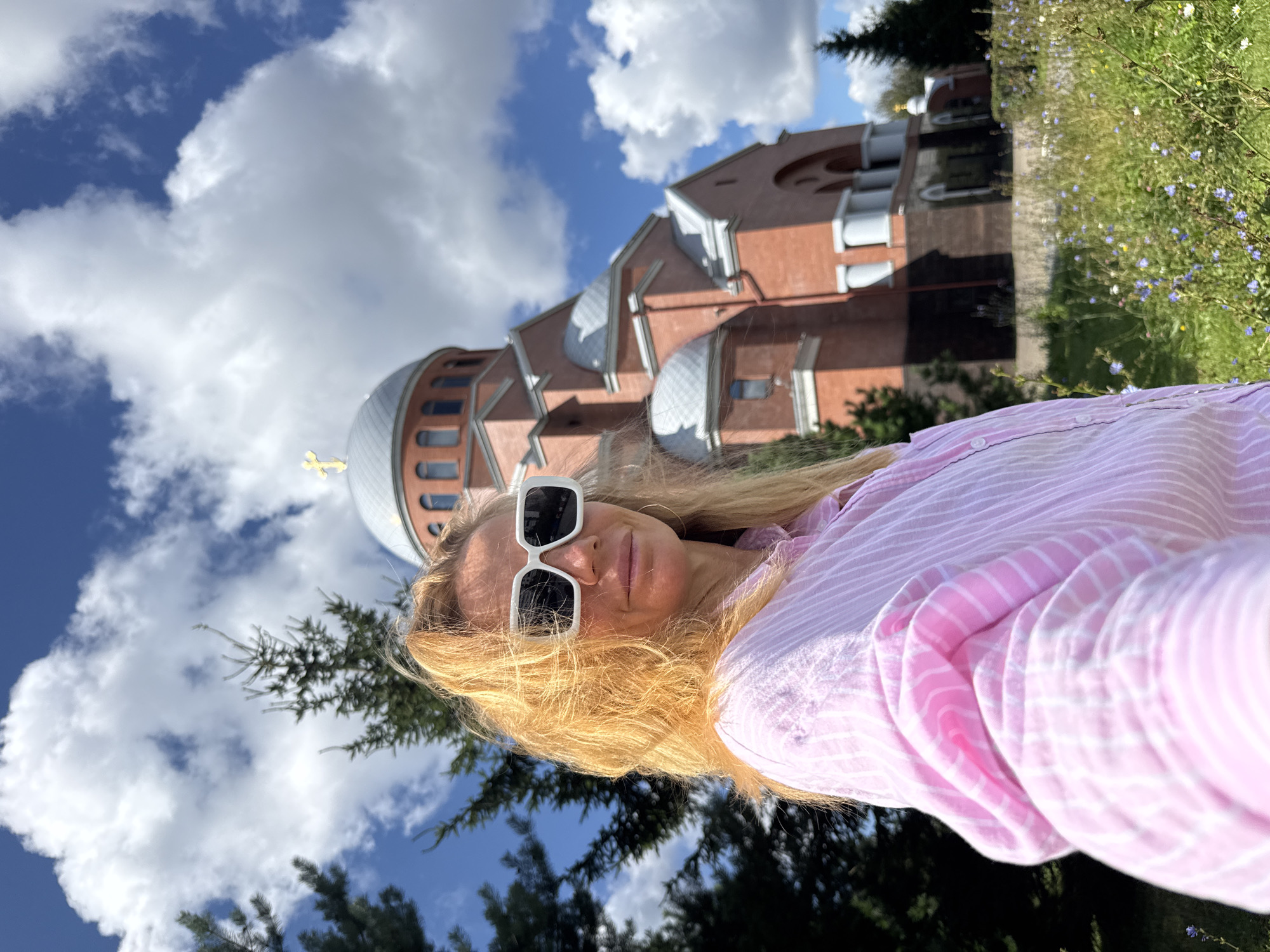 A medium shot selfie shows a blonde woman with light-colored hair, wearing white rectangular sunglasses and a pink and white striped button-down shirt. She is outdoors, with a bright blue sky and fluffy white clouds above. Behind her, a large, red brick church with a grey dome topped with a golden cross is visible. Evergreen trees are on either side of the woman, and a grassy area with small blue and yellow flowers is in the foreground. The woman is looking towards the camera, and her left arm is outstretched, holding the camera. The church appears to be the Church of the Transfiguration in Saint Petersburg, Russia.