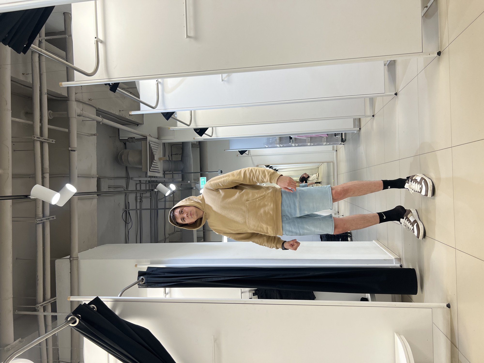 A young man stands upright in what appears to be a retail changing room or fitting area in Izhevsk, Russia. He is the central subject, looking directly at the camera with a neutral expression, his arms bent slightly with hands loosely fisted.
He is dressed in casual attire, featuring a beige hooded sweatshirt with a front pocket, light-wash denim shorts, black socks with a prominent white Nike swoosh, and white and brown sneakers.
The setting is characterized by a series of white, floor-to-ceiling partitions creating multiple individual changing stalls. Black privacy curtains are hung on silver metal rods in the foreground and mid-ground. The ceiling is an exposed industrial style, revealing white pipes, electrical conduits, and numerous white track lighting fixtures that illuminate the space brightly. Square, light-colored tiles cover the floor.
In the background, beyond the partitions, a large mirror reflects a partial view of the store, including faint shapes of other individuals. A green exit sign with a white running man symbol is visible high on a distant wall.
The scene captures a moment likely related to trying on clothes or showcasing an outfit within a modern, functional retail environment.