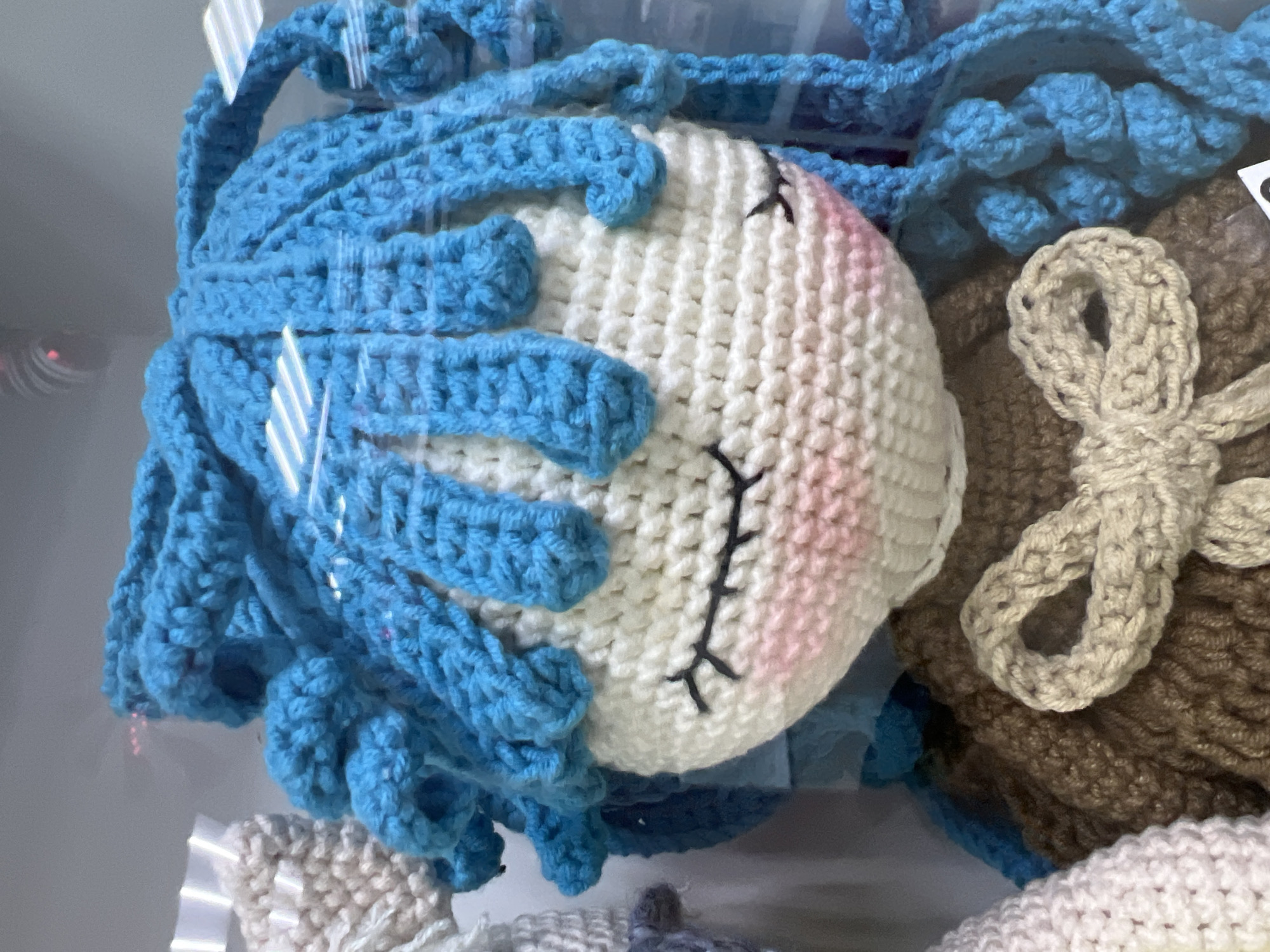 In a photograph taken in Chaikovsky, Russia, an array of handcrafted crocheted items is displayed. The primary focus is a round, white crocheted head with pink cheeks and embroidered black eyes. Adorning the head are curls of light blue yarn. Adjacent to this is a brown crocheted object with a light beige crocheted bow. The items are placed within a glass-enclosed display, possibly in a retail or exhibition setting. The environment suggests a commercial space, with the crocheted items serving as artisanal or decorative objects.