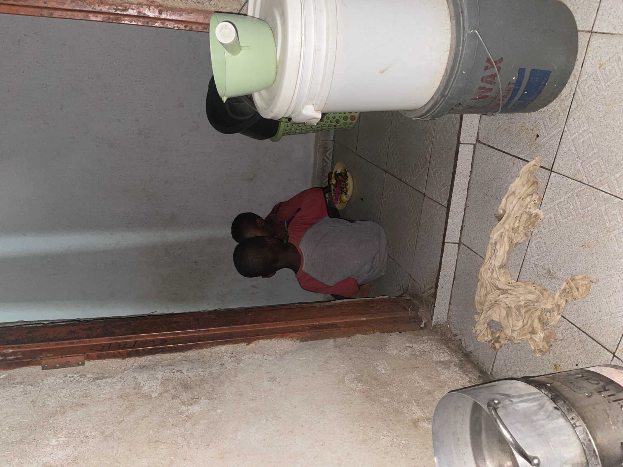 In Rido, Nigeria, the image shows two children eating from a plate on the floor. A gray and white paint bucket, a green bucket with a spout, and a green basket are stacked above them. A dirty cloth is on the tiled floor to their right. A metal pot sits on the floor in the foreground. The room has tiled floors and walls painted a light color. A brown metal doorframe is also visible.