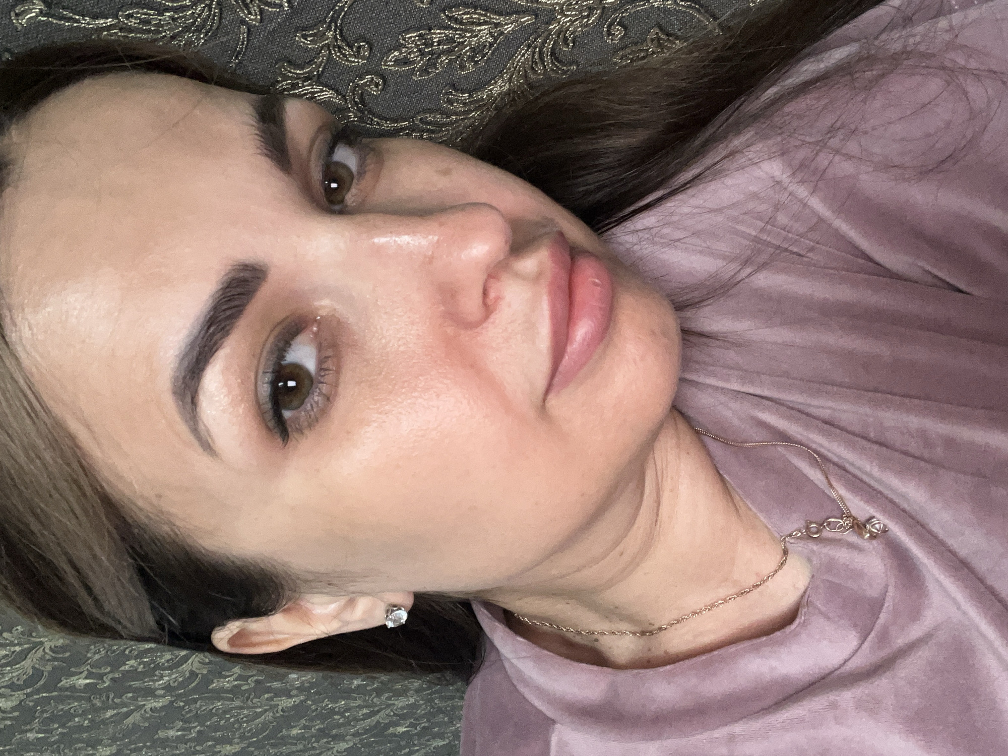 A close-up, head-and-shoulders shot shows a woman with brown hair and brown eyes, wearing a pink shirt and a gold necklace with a pendant. She is also wearing a small diamond stud earring in her left ear. The background is a textured, dark gray fabric with a gold floral pattern. Her skin appears to have some redness and oiliness.