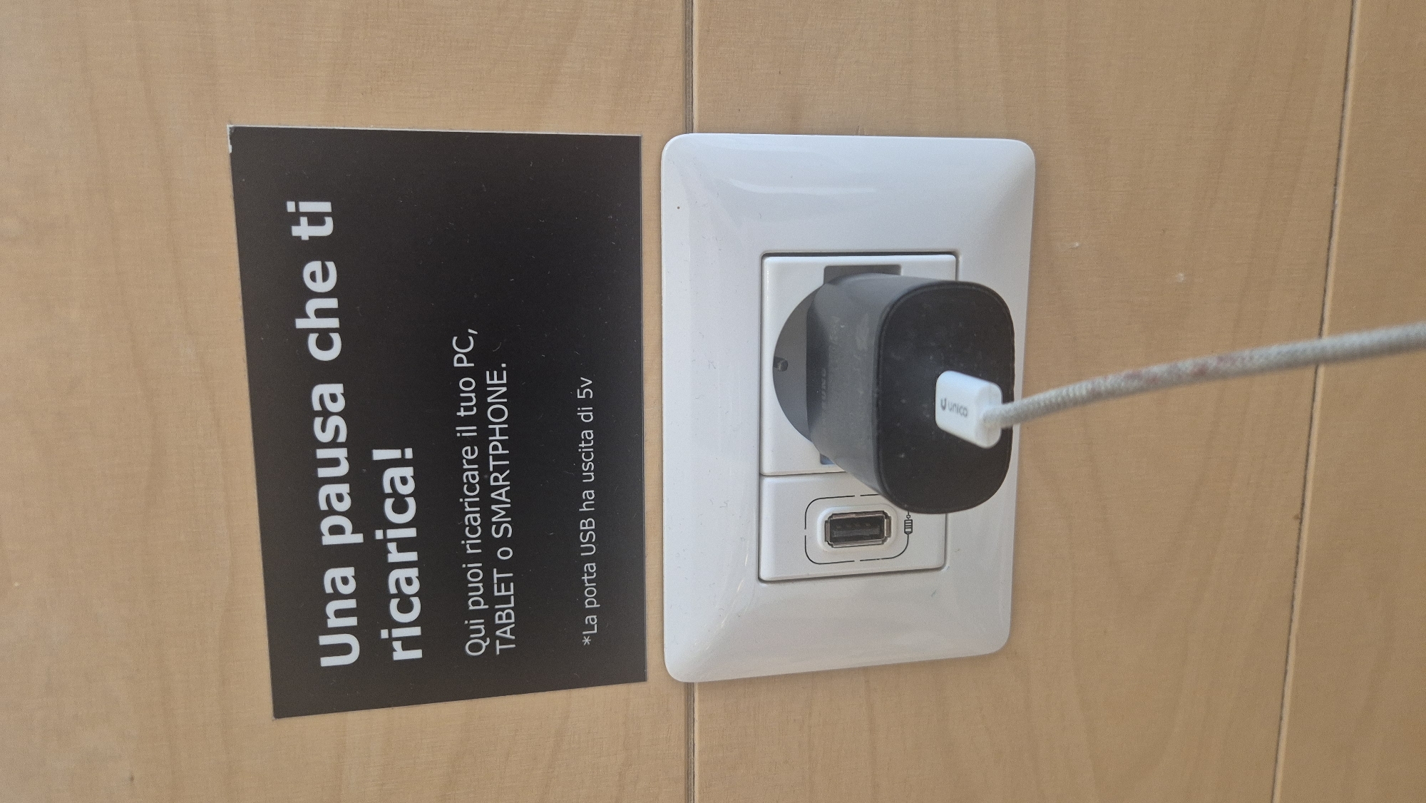 A close-up, eye-level shot shows a black power adapter plugged into a white USB wall outlet. The adapter has a white USB cable plugged into it, which trails downwards out of frame. The outlet is set into a light brown wooden wall. Above the outlet, a black sign with white text reads: "Una pausa che ti ricarica! Qui puoi ricaricare il tuo PC, TABLET o SMARTPHONE. *La porta USB ha uscita di 5v". This appears to be a public charging station, possibly located in Baronissi, Italy, as indicated by the context provided.