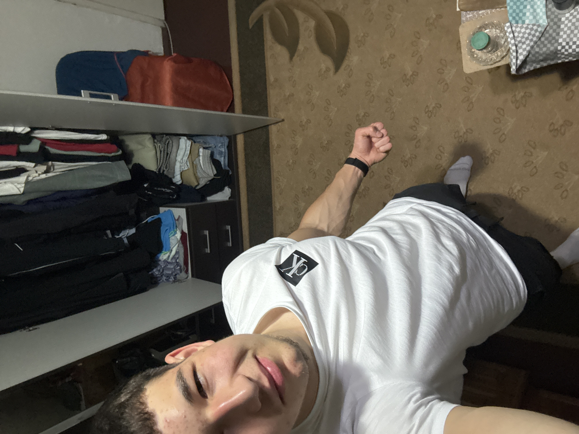 A selfie taken in what appears to be a domestic interior. A young man in a white T-shirt with a Calvin Klein logo stands, with a raised fist, partially visible at the right edge. He is wearing a black watch on his left wrist. The background to the left shows an open closet stacked with clothes and bags. Behind him is a wall and carpeted floor with a patterned design. The location is unknown but is assumed to be in Zlatograd, Bulgaria.