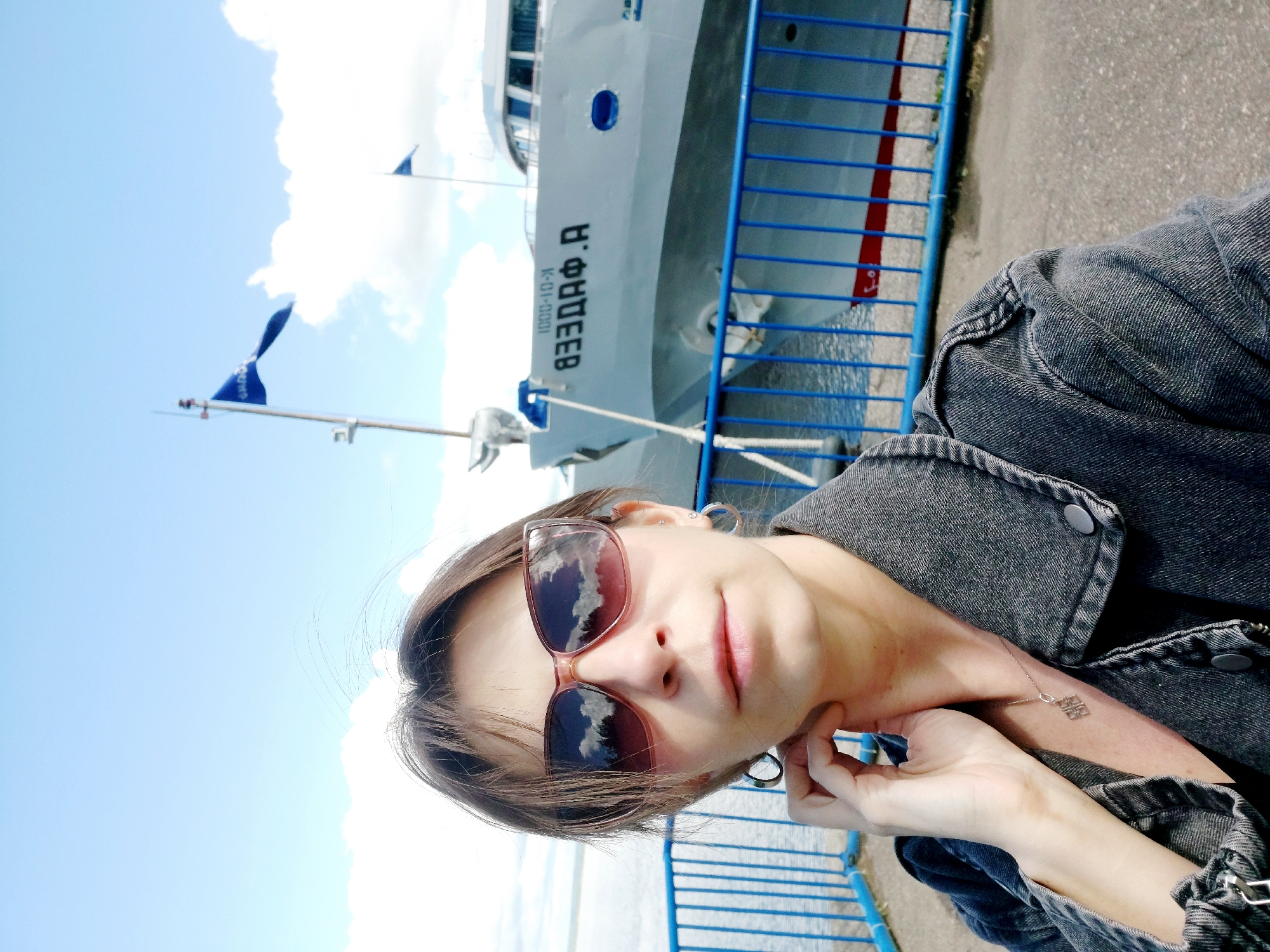 A woman takes a selfie against the background of the passenger ship "Звезда Ф.А." (Zvezda F.A., Star F.A.), registration number K-01-0001, in Chaikovsky, Russia. She wears sunglasses, a denim jacket, and earrings. The ship is docked behind a blue metal railing. The sky is partly cloudy.