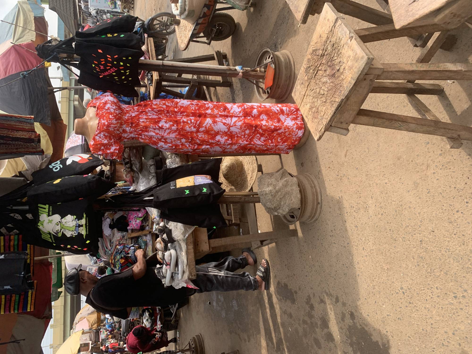 The image captures a bustling open-air market scene in Shagamu, Nigeria. In the foreground, a man wearing a black t-shirt, dark pants with white stripes, and black sandals stands behind a stall displaying an assortment of footwear. He is examining the shoes. To his right, a mannequin is dressed in a vibrant red and white floral print dress, with several black tote bags hanging behind it. The market is filled with various goods, including clothing items, bags, and other merchandise displayed on wooden stalls and tables. In the background, more stalls and people are visible, along with a motorcycle and a wheelbarrow laden with sacks. The ground is paved with a rough, textured surface. The overall impression is one of a lively commercial hub.
