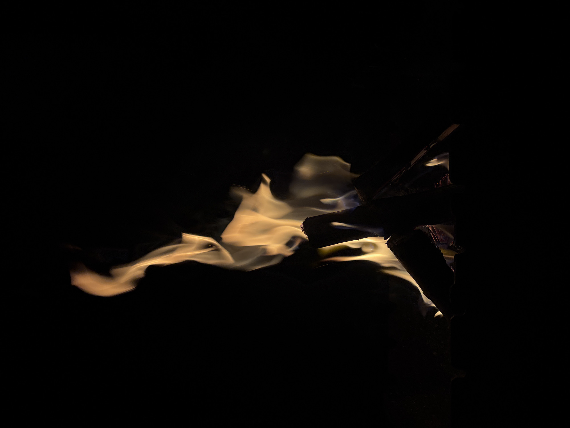 A close-up, low-angle shot captures a campfire burning in darkness. The flames are a vibrant mix of yellow and orange, dancing upwards in various shapes against a completely black background. The bottom portion of the image shows the silhouette of several logs burning, partially obscured by the intense light of the fire. The scene evokes a sense of warmth and primal energy, with the focus entirely on the dynamic nature of the flames. The location in Krasnodar, Russia, is not discernible from the image.