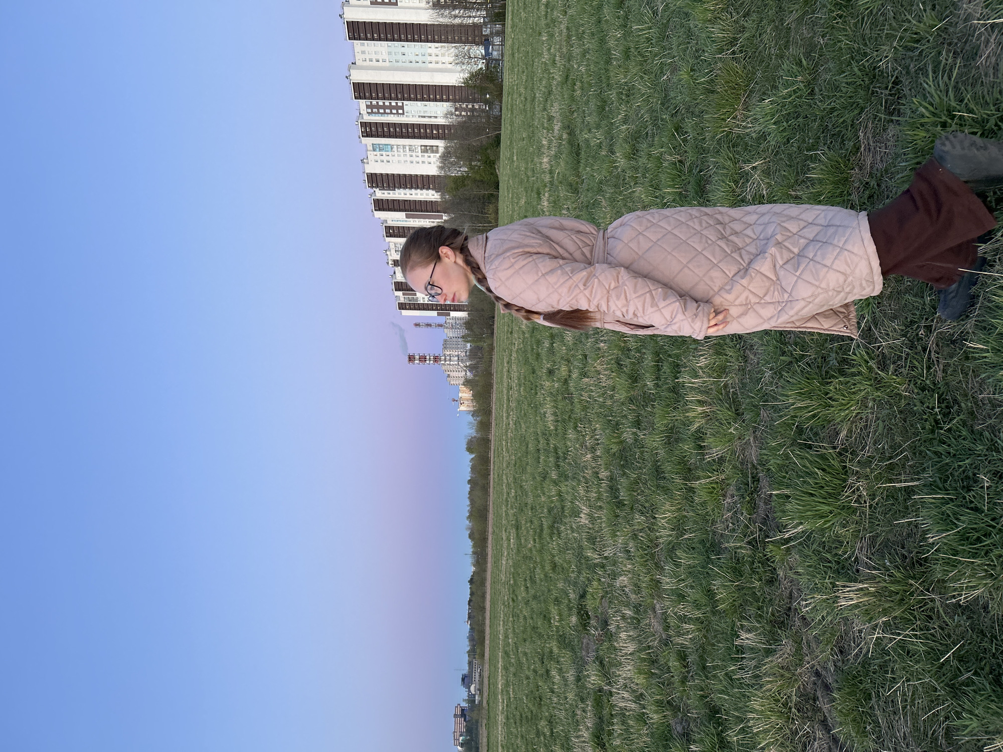 A full-length shot shows a young woman with a long braid, glasses, a quilted coat, and brown pants standing in a field of green grass. Behind her are tall apartment buildings, suggesting an urban setting. The sky transitions from a clear blue to a light purple, possibly indicating sunset or sunrise. The location is likely Saint Petersburg, Russia, given the architectural style of the buildings.