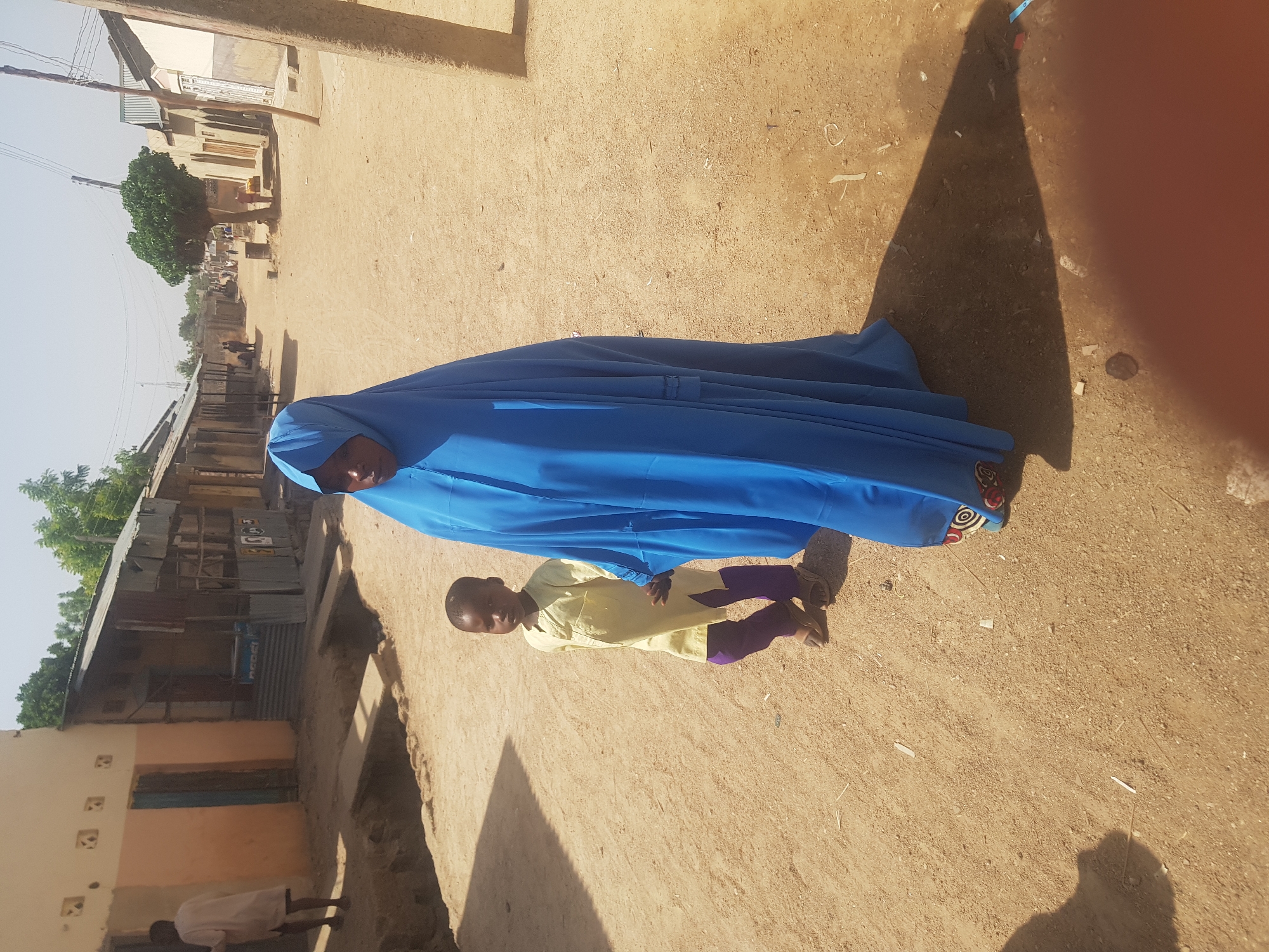 A woman in a blue full-body garment and veil walks hand-in-hand with a child on a dirt road in Dutsinma B, Nigeria. The child wears a yellow shirt and purple pants. The road is bordered by low buildings and vegetation. Shadows are cast by the sun.