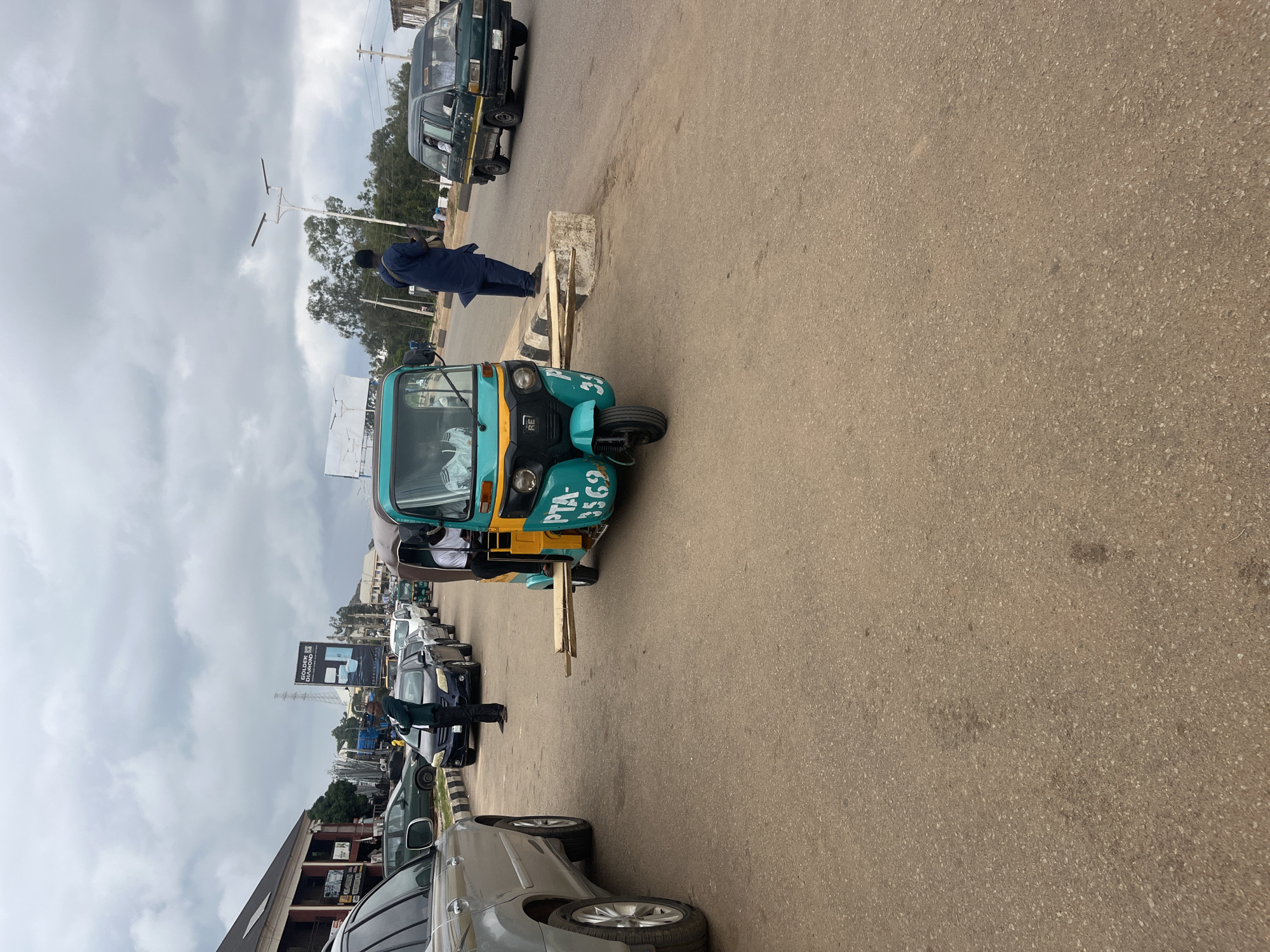 The image depicts a street scene in Gura Top, Nigeria, likely during daylight hours under an overcast sky. A teal and yellow three-wheeled auto-rickshaw, identified by the license plate "PTA-3569," is parked on the right side of the road, facing away from the viewer. Two planks of wood are extended horizontally from its right side, resting on a concrete barrier. A person, possibly the driver, is visible inside the auto-rickshaw.
In the foreground, a silver SUV is parked on the left, facing left. Beyond the auto-rickshaw, a dark-colored van is traveling away from the viewer. Further down the road, a line of cars is visible, suggesting traffic. Two men are standing in the middle of the road, one in a dark suit and the other in a dark blue robe, appearing to direct traffic. Buildings are visible on the left side of the street, and trees and utility poles line the background. The road surface is asphalt.