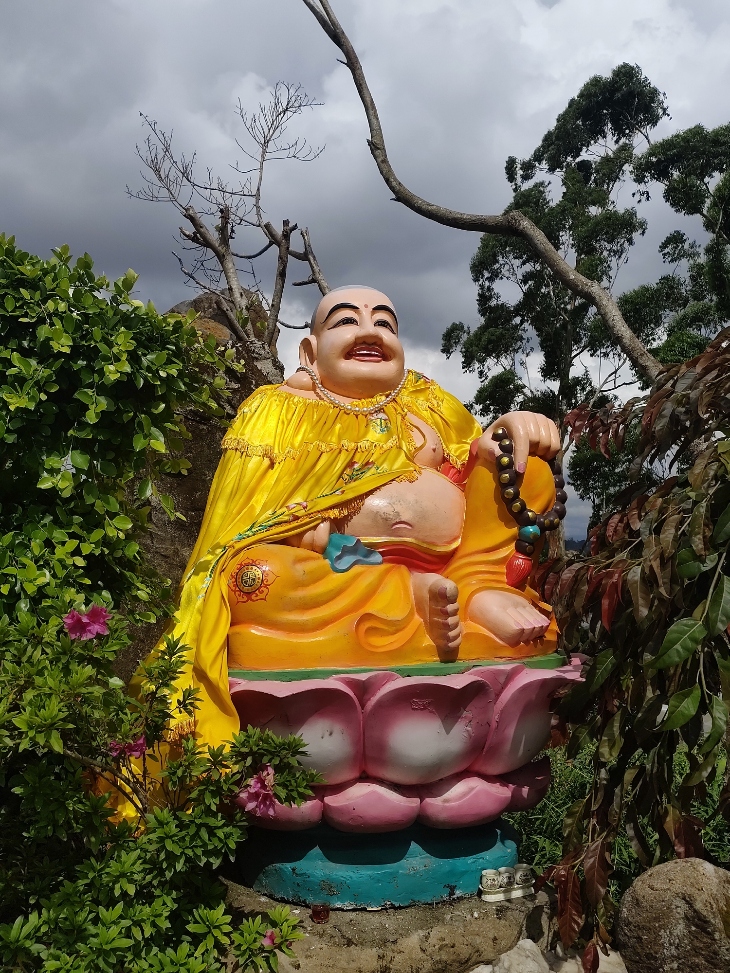 A color photograph depicts a statue of the "Happy Buddha" (Budai) sitting atop a lotus flower base. The statue is situated outdoors amid foliage and trees. The statue is dressed in a yellow robe and a necklace. The Happy Buddha holds a beaded mala in his right hand. The lotus flower base is painted with pink and white petals, with a blue and green base. The statue is surrounded by green leafy plants with pink flowers on the left side and brown-leaved foliage on the right. Behind the statue, there are bare tree branches and green trees against a sky with overcast grey clouds. Two small tea cups are set on a white tray at the base of the statue. The photograph was taken in Lâm Viên - Đà Lạt, Vietnam.