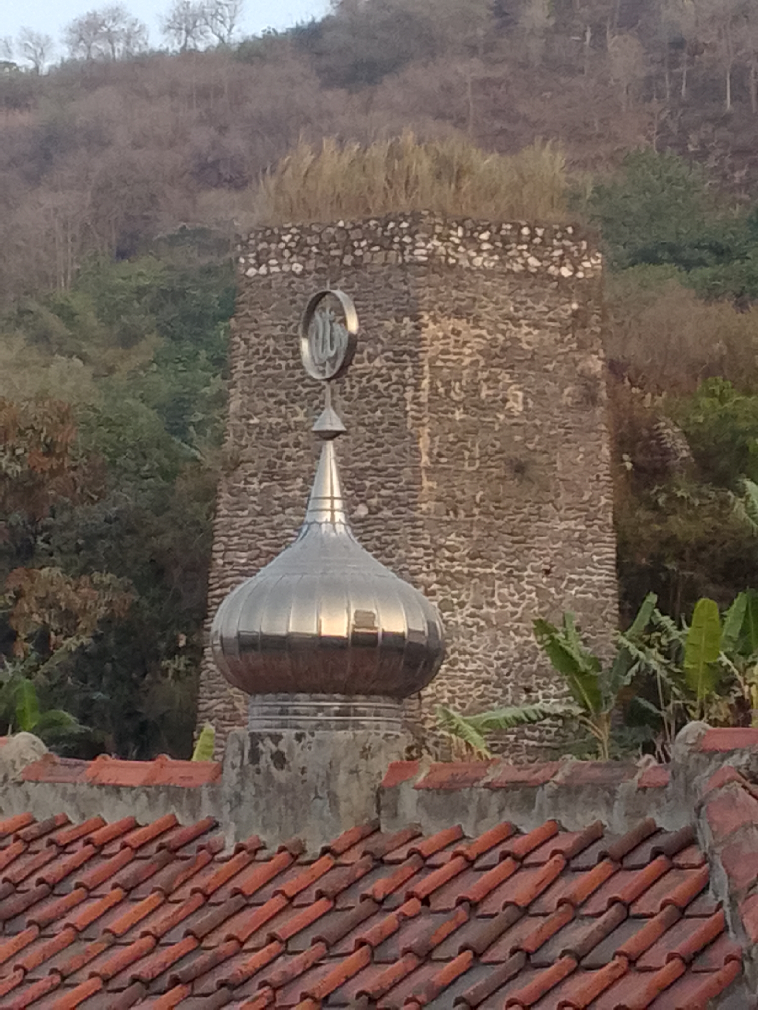 The image shows a close-up view of a mosque dome and minaret, with a stone tower in the background. The dome is silver and has a pointed finial. The minaret is also silver and has Islamic calligraphy on it. The stone tower is made of rough-hewn stones and has a weathered appearance. In the foreground, there is a tiled roof. The background consists of a hillside covered in trees and vegetation. The sky is overcast. The overall impression is one of a serene and traditional religious site. The specific location in Gunungmasigit, Indonesia is not evident from the image itself, but it is stated in the prompt.