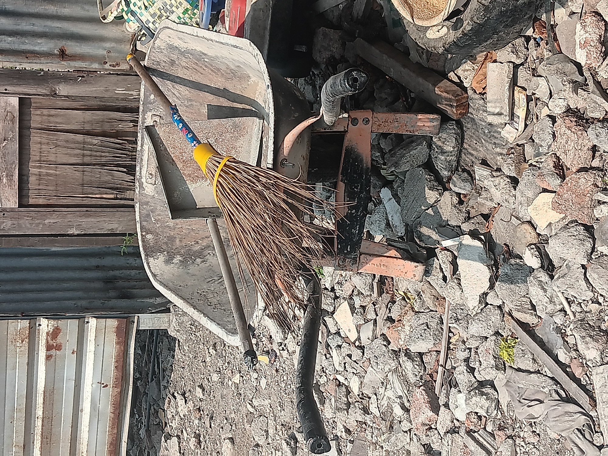 The image shows a cluttered outdoor scene with construction debris. A wheelbarrow, rusted and worn, is the central object. A broom with a yellow handle and natural bristles rests on top of the wheelbarrow. The background consists of corrugated metal sheeting and weathered wooden planks, suggesting a dilapidated structure. The ground is covered in a mixture of gravel, broken concrete, and other debris. The overall impression is one of disuse and disrepair. The location is identified as Batursari, Indonesia. There are no people visible in the image.