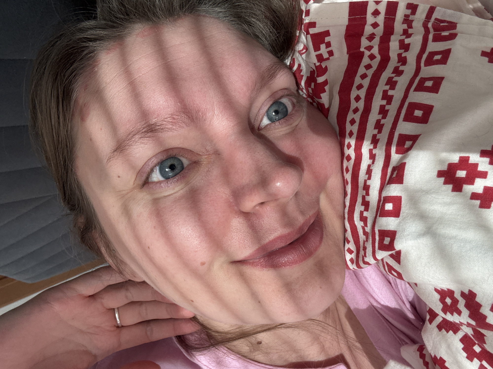 A close-up, brightly lit photograph captures a person, likely a woman, lying down with their head resting on a patterned pillow. The pillow has a white background with red geometric patterns. The person has fair skin, light brown hair, and blue eyes. Shadows from what appear to be blinds or slats are cast across their face. They are wearing a light pink top. A silver ring is visible on their left ring finger, which is touching their neck. The background is a plain, dark grey surface, possibly a couch or headboard. The location is unknown and cannot be determined as Izhevsk, Russia from the given image.