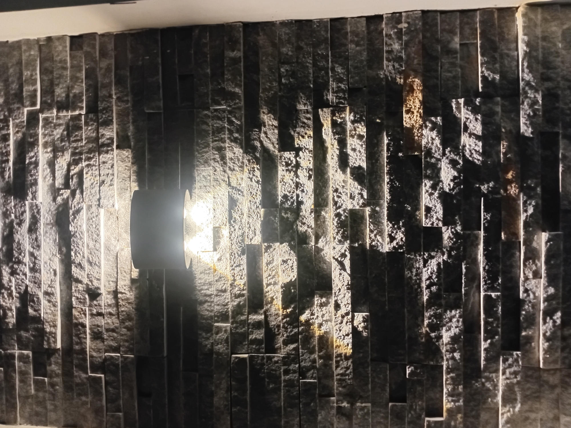 The media file shows a close-up, horizontally oriented shot of a textured, dark stone wall with a light fixture. The wall is constructed from rectangular stone tiles, laid vertically and of varying depths, creating a strong play of light and shadow. The stone appears rough and natural, with some variations in color, including hints of brown and gold amidst the dominant dark gray.
A circular light fixture is mounted on the wall, casting a bright, cool-toned light that illuminates a portion of the stone and creates dramatic shadows. The light source is intensely bright, creating a strong glare. The rest of the wall is in shadow, with subtle highlights reflecting off the textured surfaces. The top edge of the wall meets a plain, light-colored ceiling.
The scene appears to be indoors, likely in a modern architectural setting such as a hotel lobby, restaurant, or an outdoor covered area, given the material and lighting. The lighting suggests it could be nighttime or a dimly lit interior space. No people are visible in the frame. The focus is entirely on the texture and lighting of the stone wall. There is no visible text in the image. The overall mood is one of contemporary design and dramatic visual contrast.