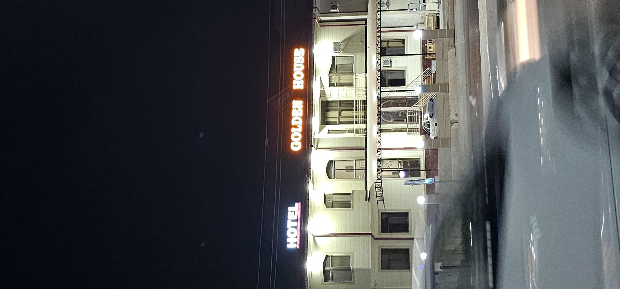 The image shows a hotel building at night, illuminated by neon signs. The signs read "HOTEL" in white and pink neon on the left, and "GOLDEN HOUSE" in orange neon on the right. The building has a light-colored facade with multiple windows and balconies. There is a covered walkway with pillars in front of the entrance. A white car is parked in front of the building. The foreground shows a blurred street or road with traffic lights reflected on the wet surface. The background is dark, indicating nighttime. The scene is located in the city of Sheyhancha, Uzbekistan.