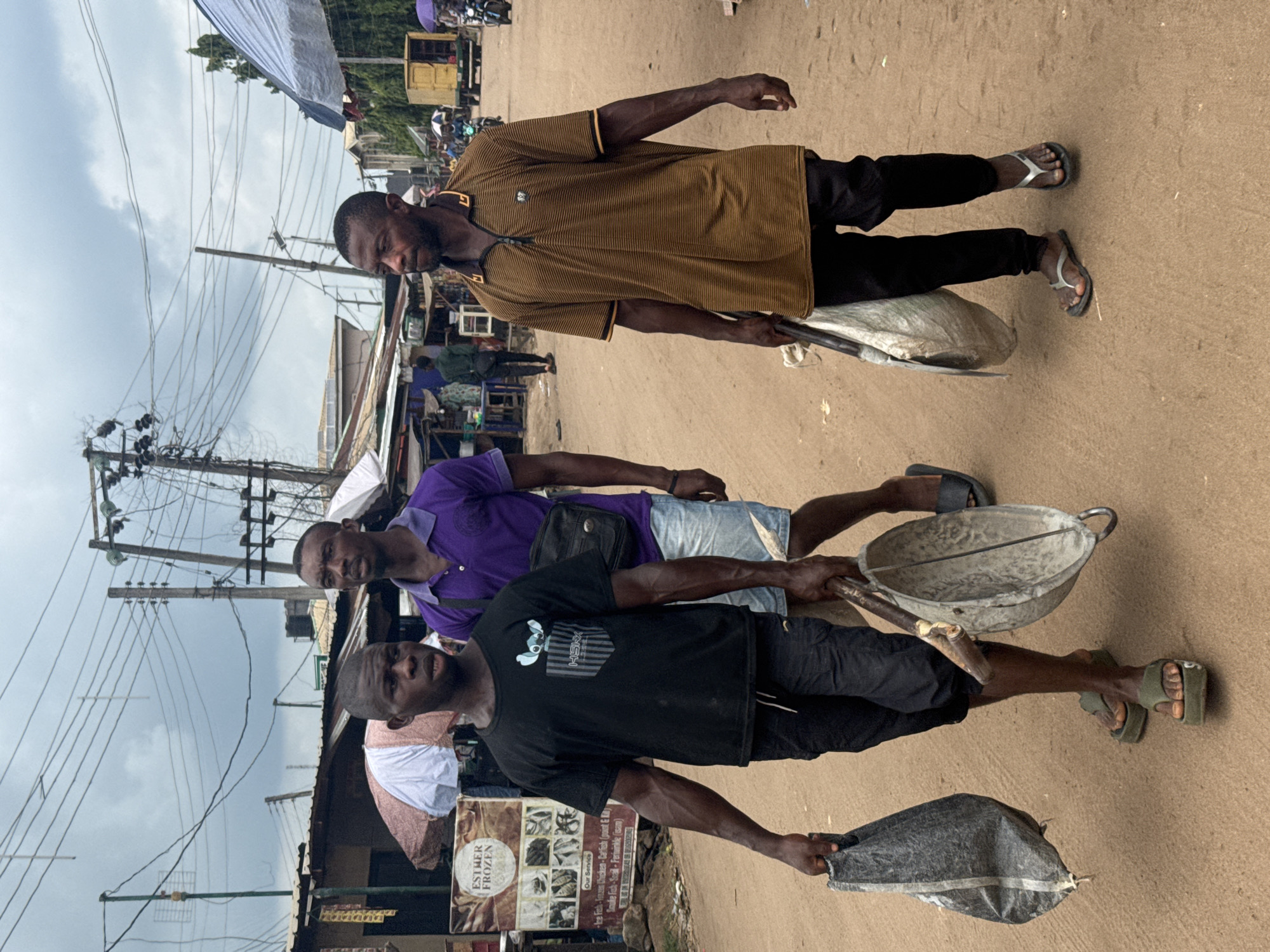 Three men are standing on a dirt road in Ihiagwa, Nigeria. Each man is carrying items, possibly for work or trade. The first man, at the top of the image, wears a brown, short-sleeved shirt and black pants, holding a filled grey sack. The second man is in a purple shirt and denim shorts, carrying a metal bucket, and has a black bag hanging from his shoulder. The third man wears a black t-shirt and black shorts and is carrying another filled grey sack. A sign reading "Esther Frozen" is visible, indicating a frozen food shop. Overhead power lines and buildings are in the background, suggesting a commercial area. The environment looks like a market or busy street with people in the background.