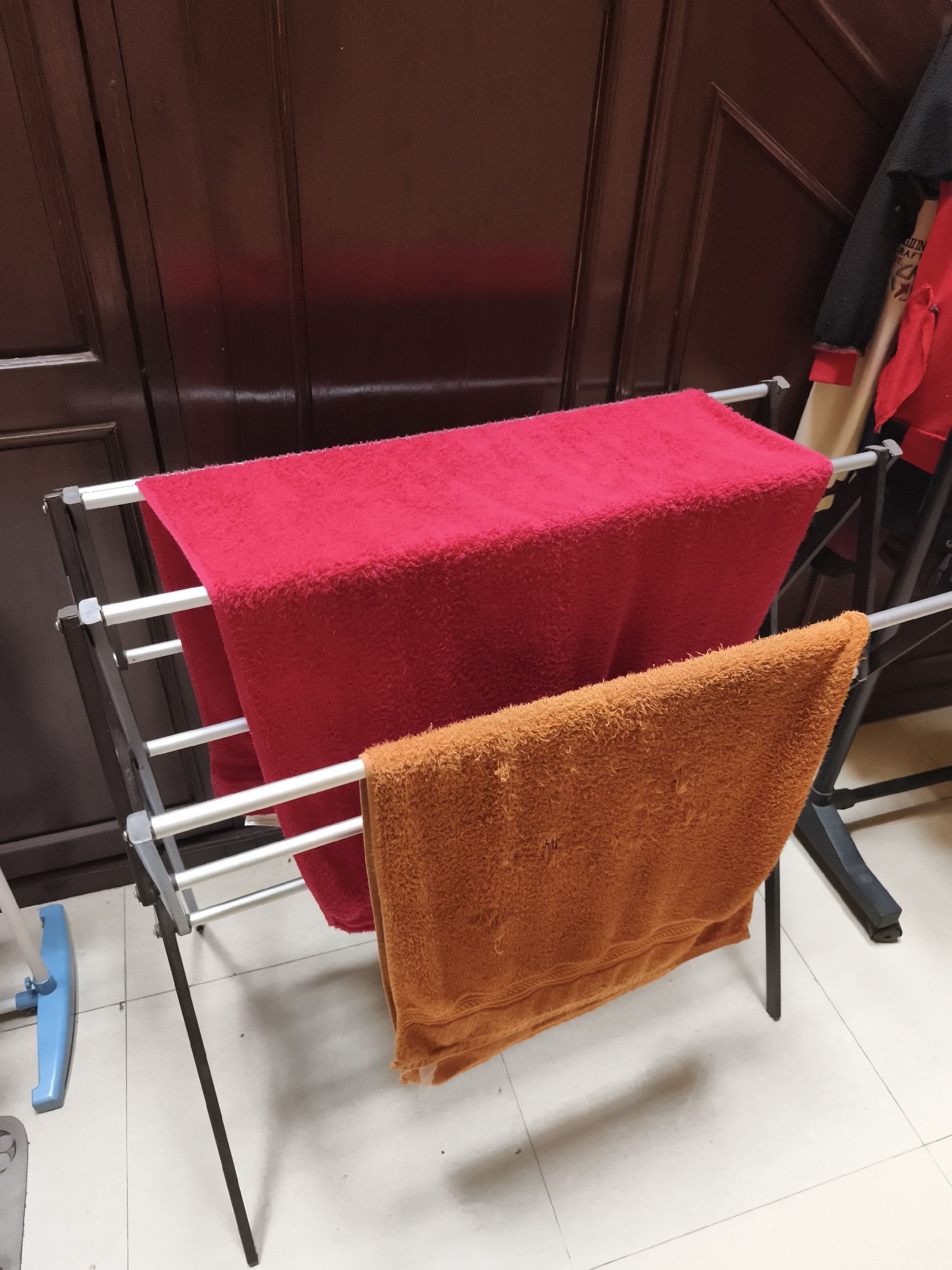 A close-up, eye-level shot shows a metal drying rack with two towels, one red and one orange, draped over its bars. The red towel is on the top bar, and the orange towel is on the bar below it. The towels appear to be made of terrycloth. The drying rack is positioned in front of a dark wooden cabinet with vertical paneling. To the left of the drying rack, a portion of a blue and white mop is visible on a light-colored tiled floor. The scene appears to be indoors, likely in a residential setting. The image does not provide any specific clues to its location in Yogyakarta, Indonesia.