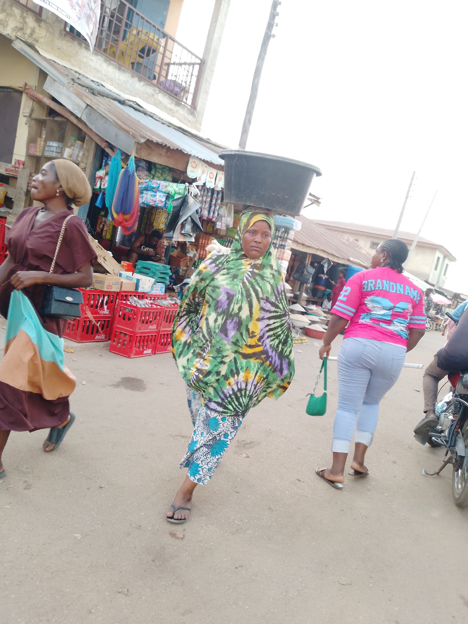 This image depicts a bustling street scene in Akwanga, Nigeria, likely during daytime, under a bright, possibly overcast, sky. The setting is an outdoor market street with vendors and pedestrians.
Three women are prominently featured. On the left, a woman in a brown dress and a light brown headscarf walks with a black shoulder bag and a two-toned light blue and tan tote bag. In the center, a woman with a colorful green, purple, and yellow patterned dress and headscarf carries a large black basin on her head, looking towards the camera with a neutral expression. On the right, a woman seen from behind wears a bright pink t-shirt with "GRANDNAM" and a light blue and white bird design, along with light blue jeans, holding a small green purse. Partial views of another person and a motorcycle are visible on the far right.
The background shows market stalls with a variety of goods, including packaged items, hanging fabrics, and red Coca-Cola crates. A person in a dark t-shirt with partially visible text, possibly "SIKOES," is seated among the crates. The buildings feature corrugated metal roofs and an upper-story balcony, suggesting a typical commercial and residential street in the region. The activity centers around daily commerce and movement through the market. The ground is an unpaved or lightly paved surface.