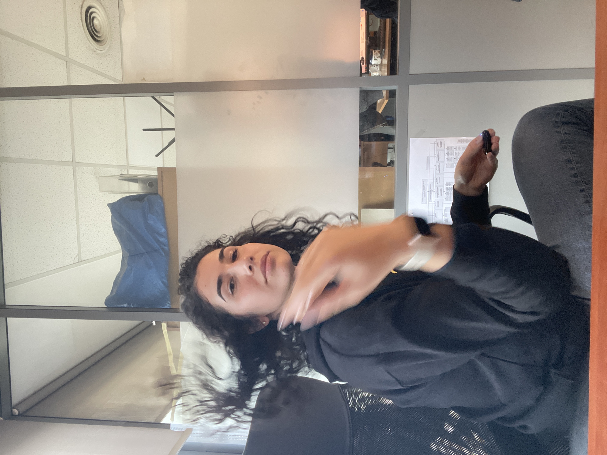 A young woman with dark, curly hair and a neutral expression is seated in an office chair, looking directly towards the camera. She is wearing a dark long-sleeved top and dark pants. Her right arm is raised and blurred, suggesting motion, while her left hand holds a small, dark object, possibly a pen or marker.
The setting is an indoor office environment, characterized by light-colored partitions that divide the space. Some partitions are opaque, while others appear to be reflective or semi-transparent, showing glimpses of objects and reflections in the background. Above, a white suspended ceiling with square tiles and a round air vent is visible. In the background, behind a partition, a blue bag or tarp-like object and a light-colored box can be seen. To the woman's right, a document or sheet of paper with printed text and diagrams is laid out on a surface, though the details are not legible.
The activity appears to be a casual moment, perhaps a brief pause from work for a photograph, with the woman's hand caught in motion. The lighting suggests an indoor daytime setting. No specific weather conditions are indicated. The provided location context is Sofia, Bulgaria, though no specific visual cues in the image confirm this. The woman's expression is calm and neutral.