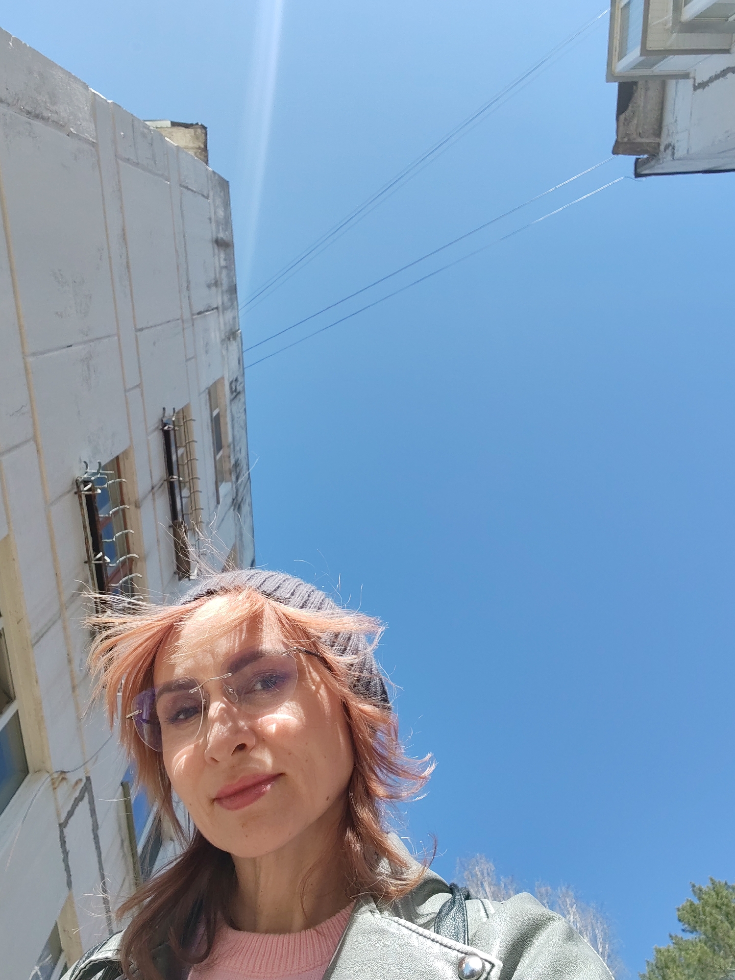 A woman with pink-tinged hair wearing a grey beanie and rimless glasses poses for a selfie. She is wearing a pink sweater and a green jacket. The background features a bright blue sky, a large white building with barred windows, and electrical wires. The photo was taken at a low angle looking up. The location is Chaikovsky, Russia.