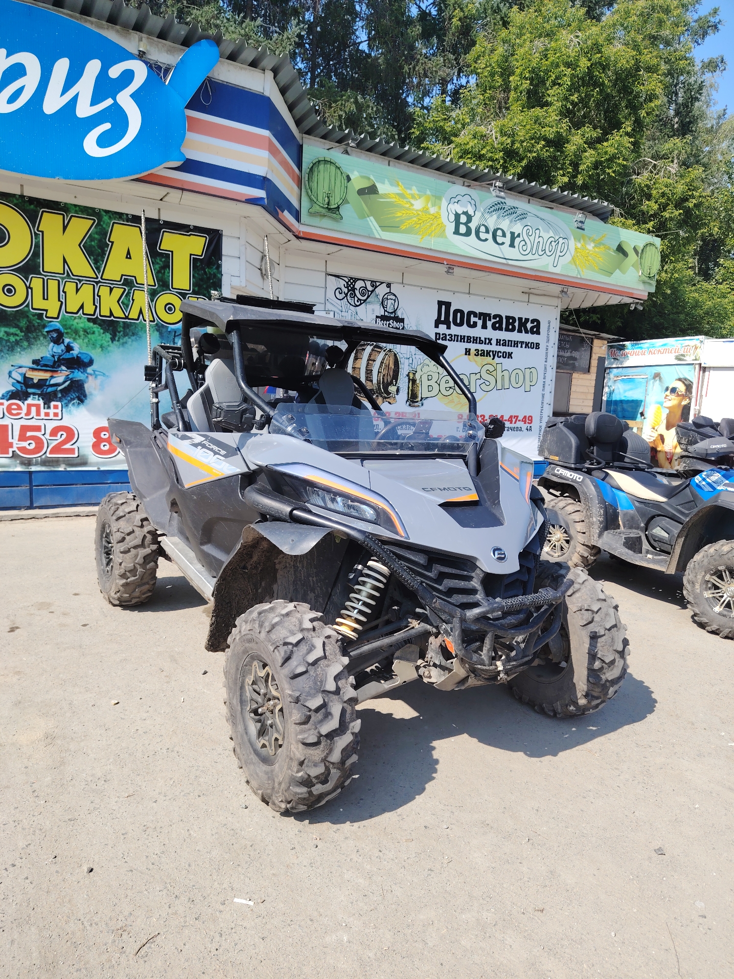 An outdoor, slightly low-angle medium shot shows two all-terrain vehicles (ATVs) parked on a paved surface in front of a building. The ATV in the foreground is gray and black with orange accents, and the ATV in the background is blue and black. Both ATVs have large, knobby tires. The building behind the ATVs has a corrugated metal roof and several signs. One sign, in large yellow lettering, says "ОКАТ" followed by "ОЦИКЛО". Another sign above it has a blue oval shape with white lettering that appears to be Cyrillic. A green sign with "BeerShop" written on it in white lettering is also visible. The scene appears to be in a commercial area with trees in the background. The lighting suggests it is daytime.