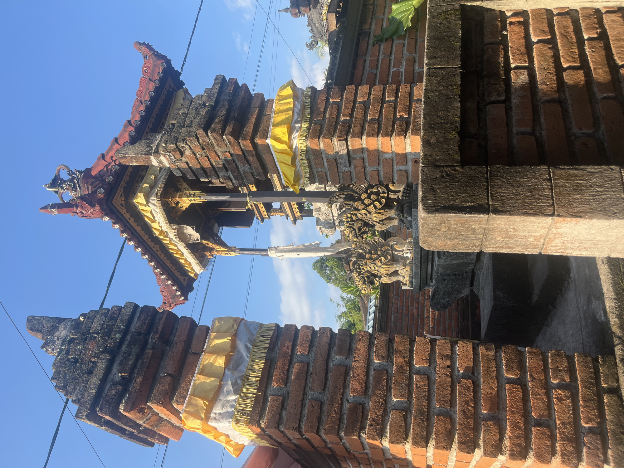 The image displays a traditional Balinese architectural scene under a clear blue sky in Mendoyo, Indonesia. The view is angled upwards, showcasing elements of a gate or shrine.
In the foreground, two vertical structures made of reddish-brown bricks are prominent. The left pillar is partially visible, topped with layers of white and yellow ceremonial cloth with a fringed edge. The right foreground features a darker, weathered brick wall capped with a dark stone slab, showing signs of wear and possibly biological growth.
The central subject is a multi-tiered shrine or altar constructed from similar reddish-brown bricks. This structure features an ornate, red-tiled roof with gracefully curved eaves and white decorative elements. Beneath the roofline, golden architectural details are visible. On the upper portion of the brick base, two intricate grey stone statues, resembling guardian figures with decorative wings and swirling motifs, are positioned. A white ceremonial banner hangs centrally from the shrine's upper section, and a yellow ceremonial umbrella is attached to its right side.
The sky is a vibrant blue with a few scattered white clouds. Thin black power lines traverse the upper half of the frame. In the mid-ground and background, glimpses of green foliage from trees and additional brick walls or structures are visible. The scene is illuminated by strong natural daylight, casting distinct shadows and highlighting the textures of the bricks and carved stonework. No people are present in the image.