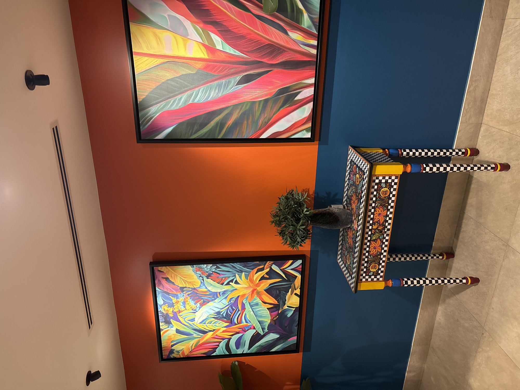 The image shows a brightly decorated hallway, likely in Amman, Jordan, featuring two vibrant abstract paintings of tropical foliage. The walls are painted in contrasting colors: the upper half is a warm, orangey-red, and the lower half is a deep blue.
On the orange wall, to the left, hangs a framed painting filled with lush green leaves, yellow and orange flowers, and touches of red and purple. To the right, another similarly framed painting displays large, red and pink leaves with yellow accents.
In front of the blue wall, positioned between the two paintings, is a narrow, decorative console table. The tabletop is adorned with a black and white checkerboard pattern, interspersed with floral motifs in orange and yellow. The table has four distinctively decorated legs, also featuring black and white checkerboard patterns, along with bands of blue and red.
On the table sits a dark, textured vase containing a small arrangement of green foliage. The floor is tiled with light-colored, square tiles. Above, the ceiling is off-white, with a dark vent running horizontally and a small, black spotlight angled downwards. The lighting in the scene is warm, casting subtle shadows and highlighting the textures and colors of the artwork and furniture. There are no people present in the image.
