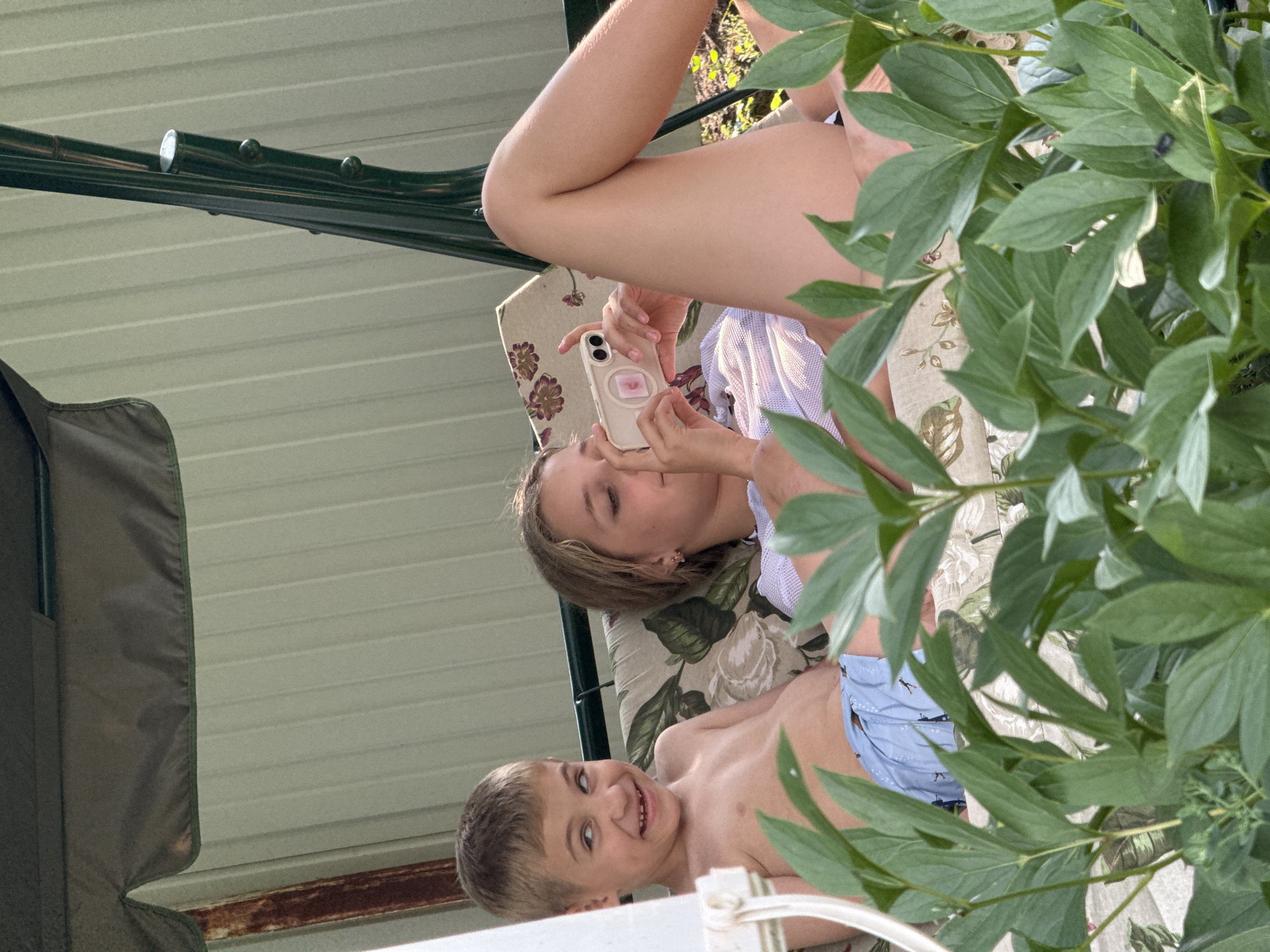 In Nizhne-Chufichevo, Russia, two children are seen relaxing on a swing set. A young boy, shirtless and wearing light blue patterned swim trunks, sits on the left, making a playful facial expression. To his right, a girl, wearing a white lacy top, is lying down and holding a smartphone, seemingly taking a photo or video. The swing set has a dark green frame and a muted patterned cushion. The background consists of a light-colored, ribbed siding. In the foreground, lush green foliage obscures a portion of the scene, hinting at an outdoor, possibly garden, setting. The overall lighting suggests it is daytime.