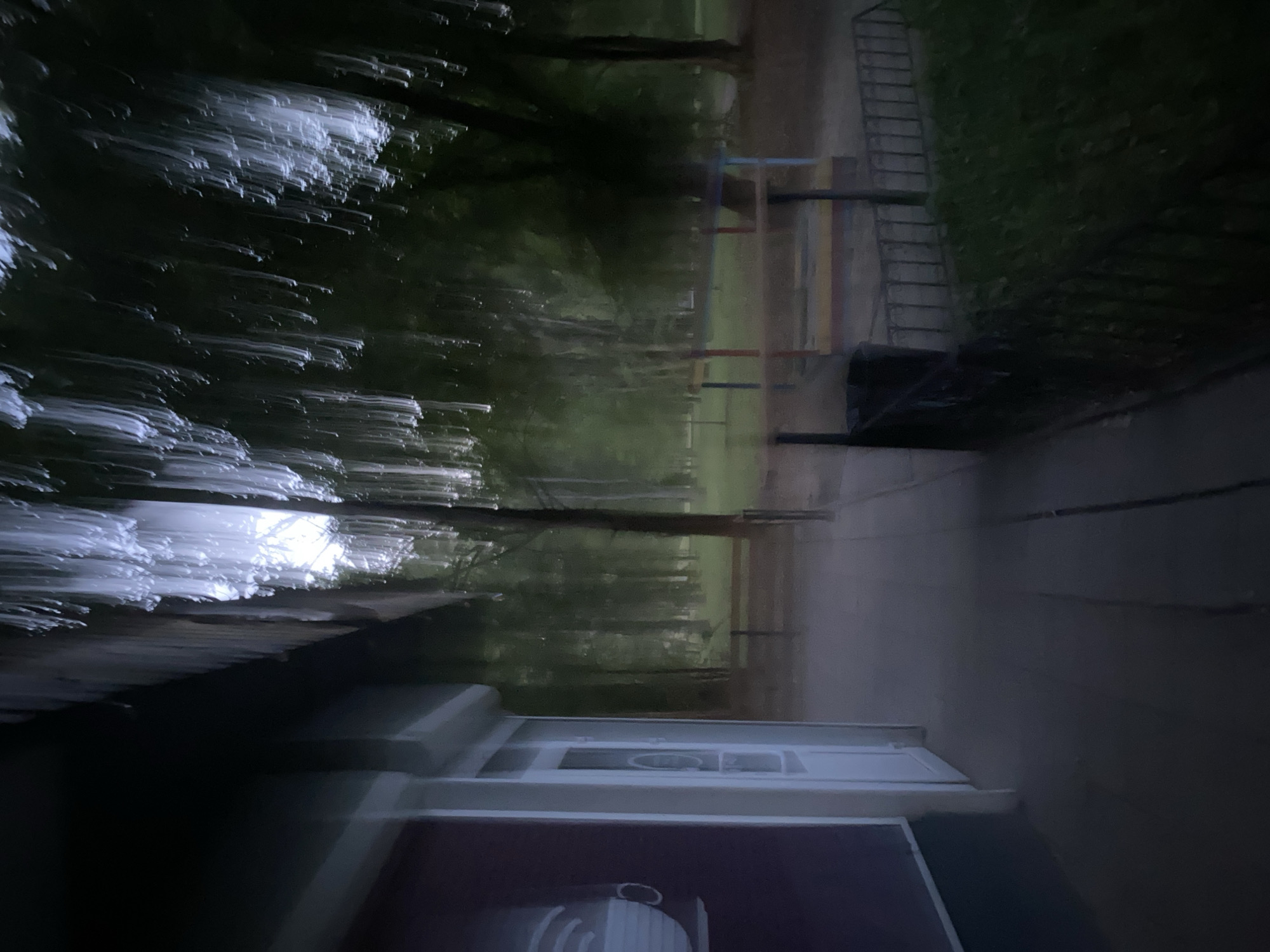 This is a night time photograph taken outdoors in Poltava, Ukraine. The image is blurred due to motion, giving a sense of movement or a low light setting. In the foreground to the left, there is the side of a building with a dark maroon colored wall and a white door. The building has an overhang that casts a shadow. To the right of the building is a sidewalk. Beyond the sidewalk, there is a grassy area with a low black metal fence. In the background, there is a park or wooded area with trees. There is some playground equipment visible in the background, including a structure with red and blue supports and a yellow platform. The lighting appears to be from streetlights or the sky, creating streaks of white light in the upper portion of the image, possibly indicating rainfall or movement of foliage.