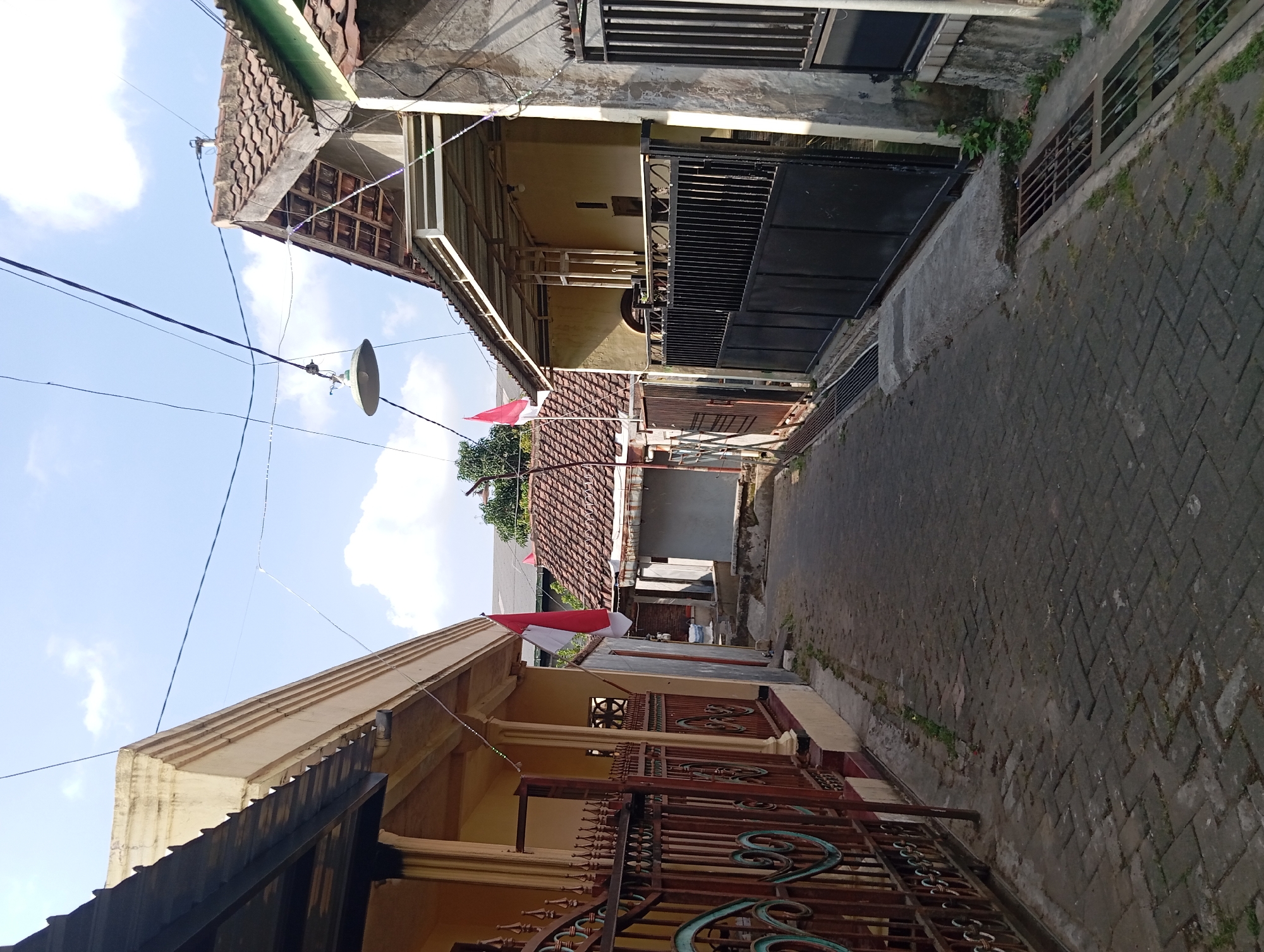 This image captures a narrow, paved alleyway in Nogosari, Indonesia. The alley is flanked by residential buildings with tiled roofs. Two Indonesian flags are prominently displayed, one on the left side and another further down the alley. An overhead lamp hangs from wires stretched across the alley. The pathway is constructed of dark, interlocking bricks with some areas of grass growing between them. Drainage grates are visible along the sides of the alley. The sky is partly cloudy.