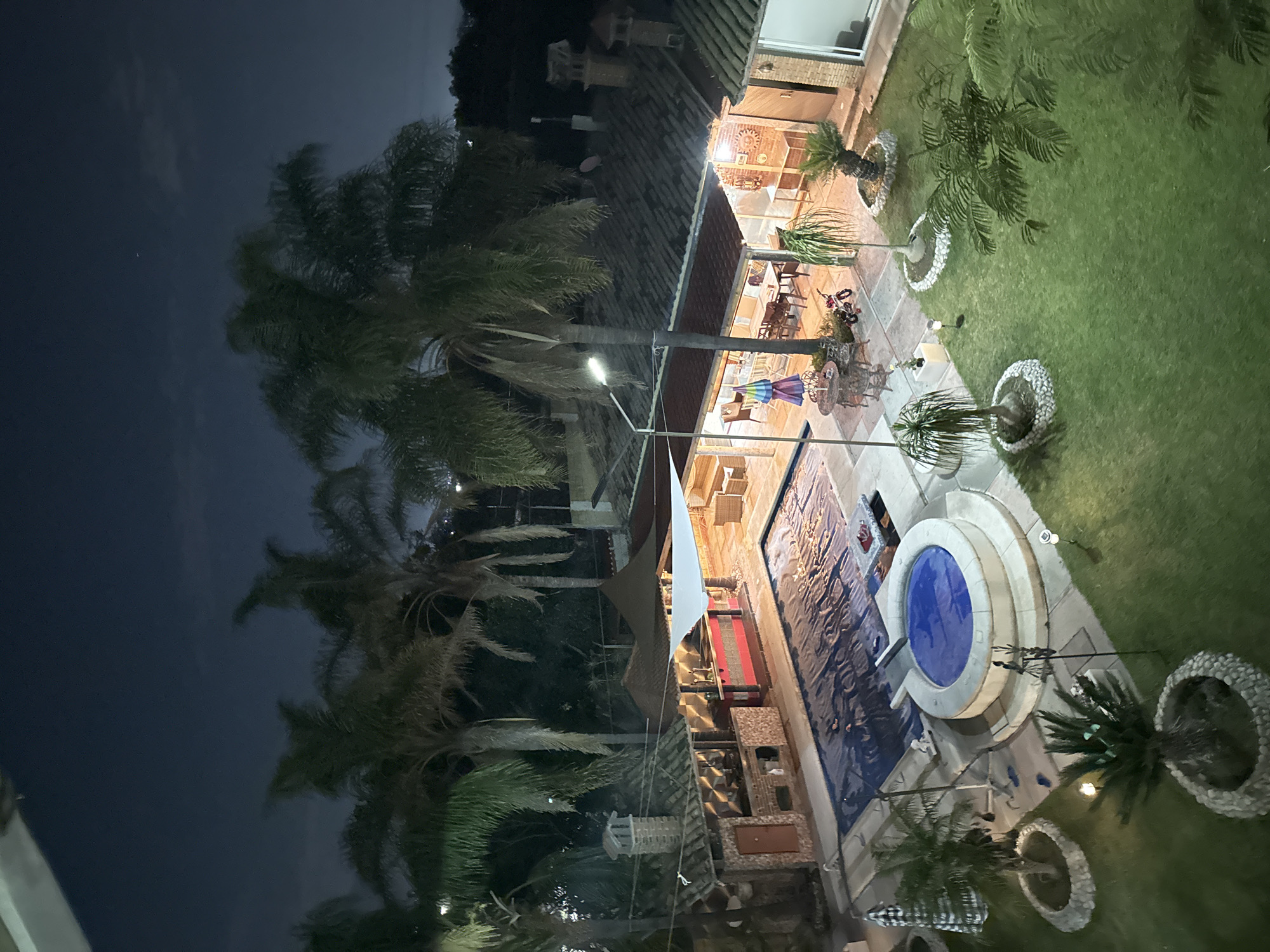 An aerial view depicts a residential backyard in Heroica Puebla de Zaragoza, Mexico, at night. The area is divided into a dark, tree-dominated zone on the left and an illuminated area on the right.
The illuminated section features a house with a tiled roof, outdoor furniture, including tables and chairs under a partially visible parasol. A swimming pool with a circular jacuzzi is present, alongside neatly manicured lawn areas interspersed with palm trees and decorative stone arrangements.
Artificial lighting illuminates the entire scene. A street lamp is visible in the background among the palm trees.