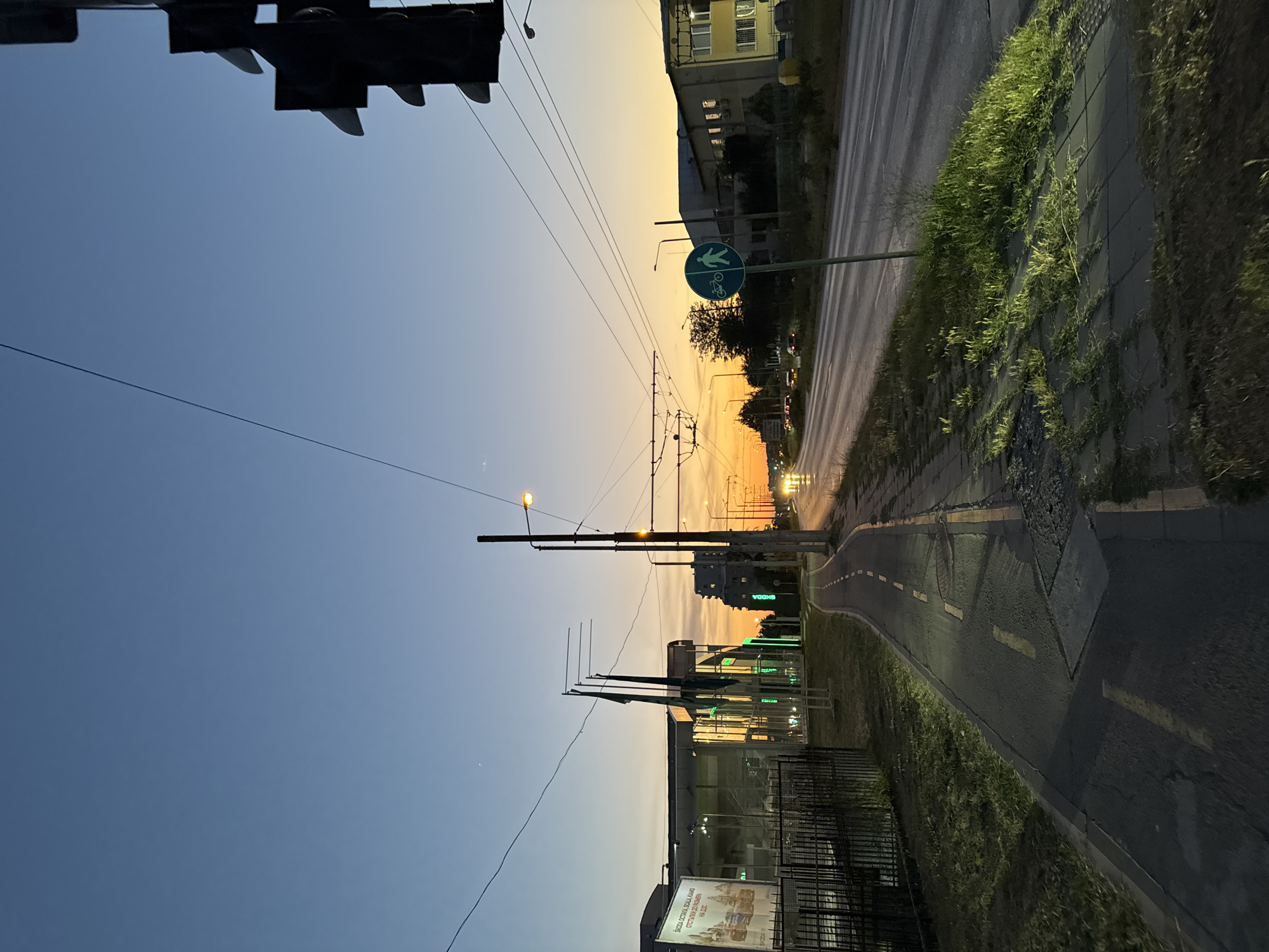 The image depicts an urban street scene in Varna, Bulgaria at dusk. A road with traffic lights and overhead wires stretches into the distance, illuminated by the fading light of the setting sun. A dedicated bicycle lane runs alongside the road, bordered by a sidewalk and patches of grass. Buildings, some with illuminated windows, line the street on both sides. Traffic lights are visible, as well as a pedestrian and bicycle crossing sign. The sky is a gradient of blue and orange, indicating the transition from day to night. No people are visible in the image.