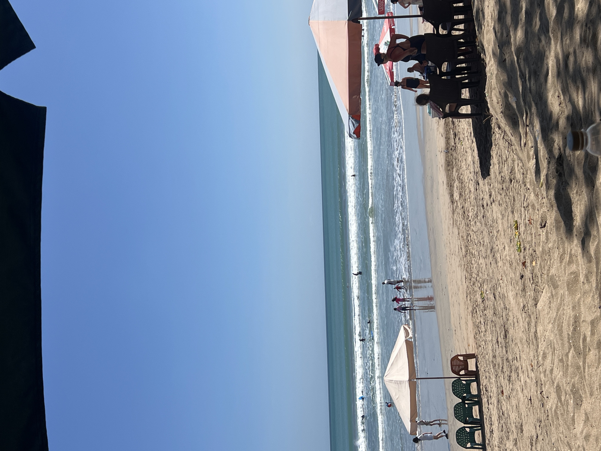 A beach scene in Kuta, Indonesia, under a clear blue sky. In the foreground, a sandy beach with some scattered leaves and debris. There are several plastic chairs set up on the sand, some under a large umbrella. A person is partially visible in the lower right corner, and a water bottle is in the extreme foreground.
In the middle ground, the ocean meets the shore with gentle waves. Several people are in the water, some appearing to be surfing or learning to surf. There are more umbrellas and chairs set up closer to the water's edge. A group of children and adults are standing in the shallow water near one of the umbrellas.
In the background, the ocean stretches out to the horizon, with more surfers visible in the distance. The overall atmosphere is one of a sunny, active beach day.