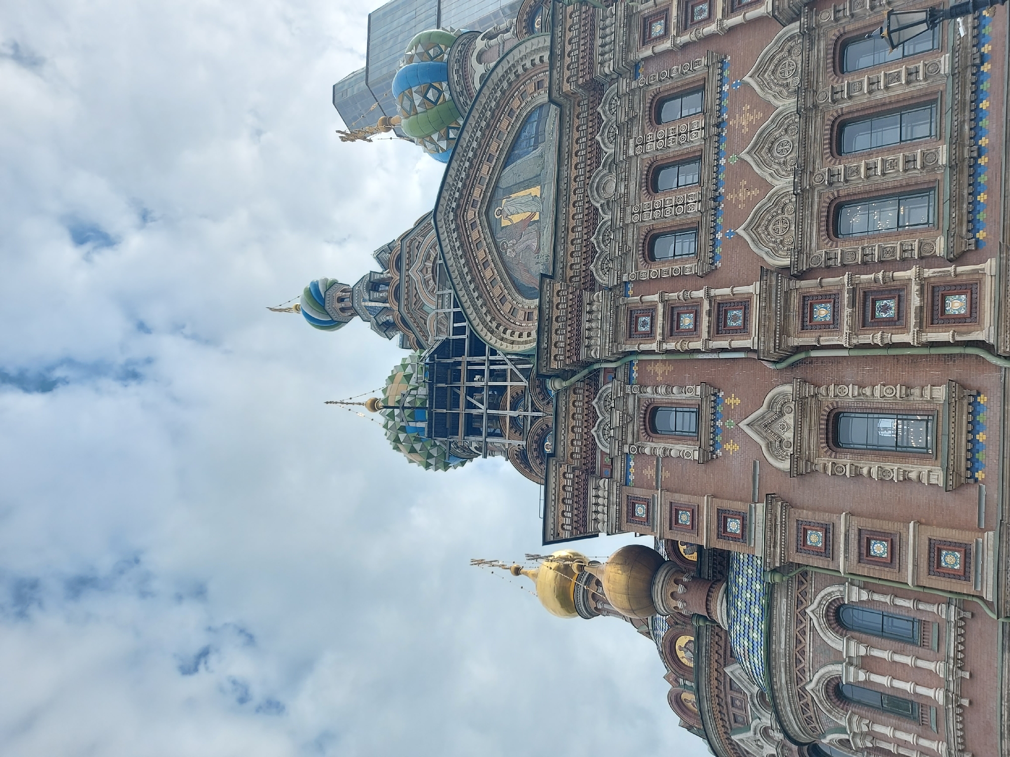 The image shows the Church of the Savior on Spilled Blood in Saint Petersburg, Russia. The building is ornately decorated with colorful domes, intricate brickwork, and mosaics. Scaffolding is present around one of the domes, indicating ongoing restoration work. The sky is cloudy, and a modern glass building is visible in the background, contrasting with the historic architecture of the church. There are no people visible in the scene.