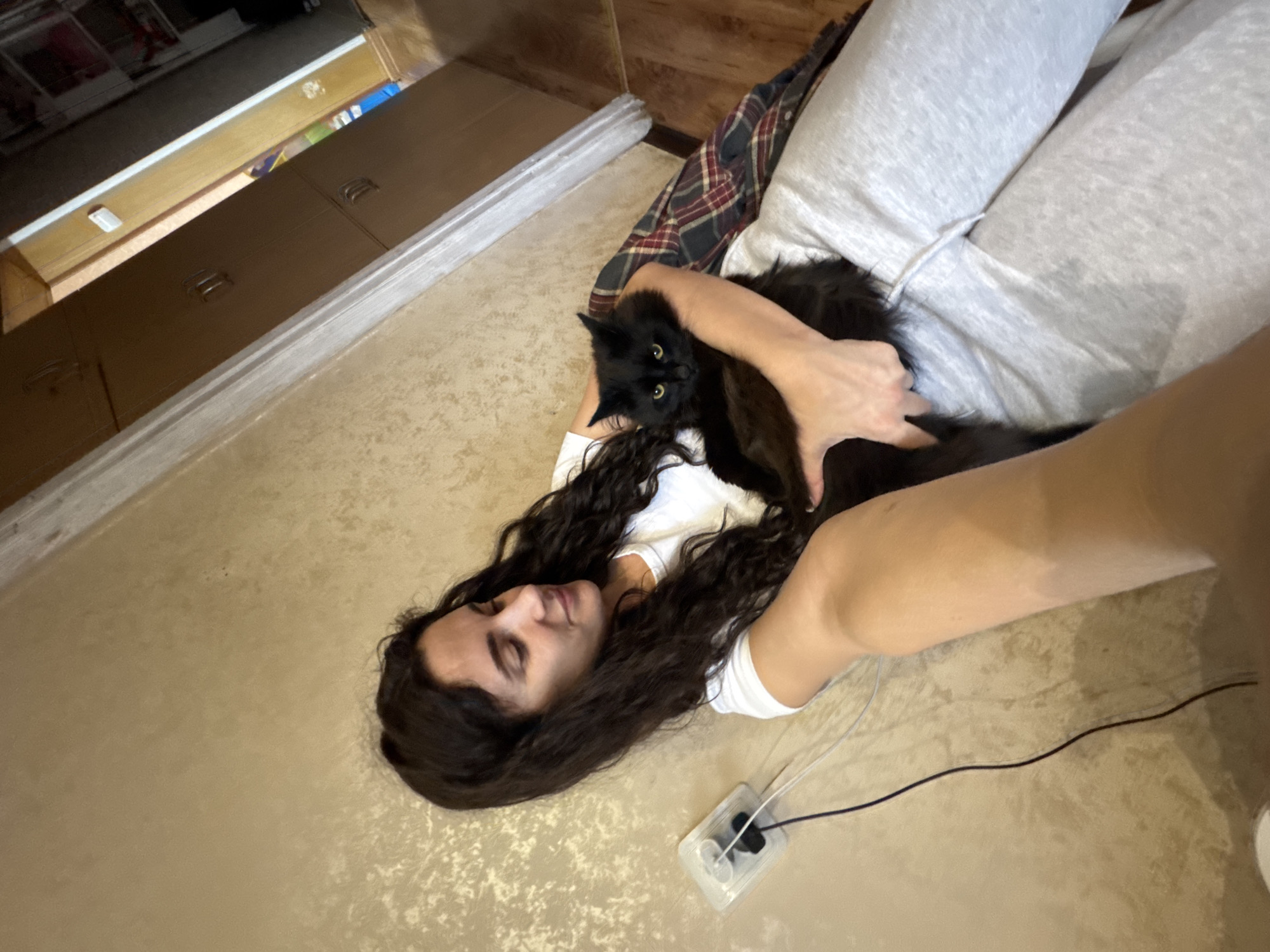 A woman with long, dark, curly hair is lying on a beige surface, possibly a floor or bed, and holding a black cat. The woman is wearing a white shirt. She has dark eyebrows. The cat is looking directly at the camera. Part of the woman's leg is visible, covered in grey sweatpants. There is a black and white power adapter with a cord next to the woman. A piece of plaid fabric is visible in the background. Furniture is visible in the background, including what appears to be a cabinet or dresser.
There is no information in the image that would indicate that it was taken in Chaikovsky, Russia.
