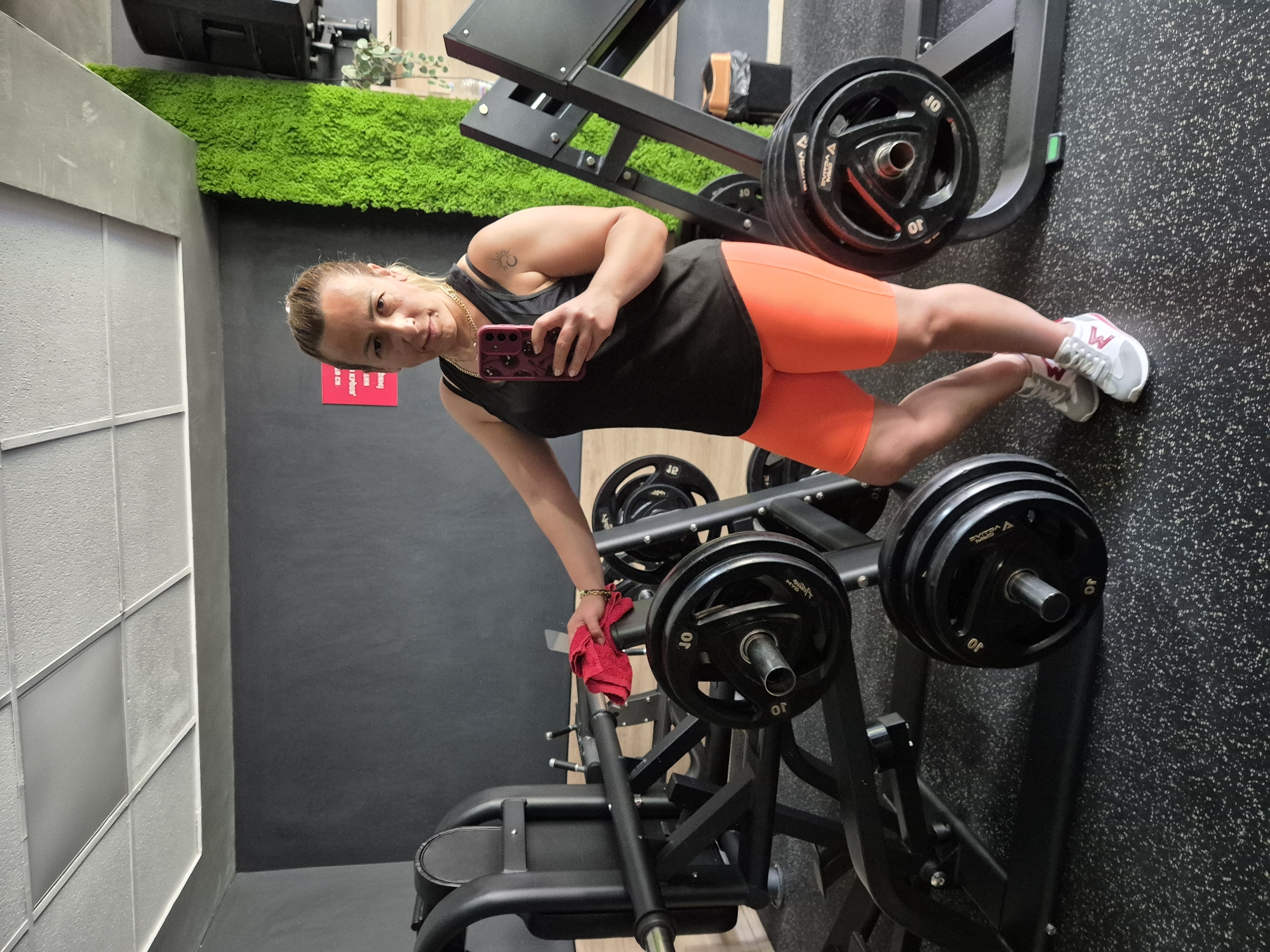A woman in athletic wear takes a selfie in a gym. She has blonde hair pulled back in a ponytail, a black tank top, and bright orange athletic shorts. She is holding a red cloth in her left hand and her phone in her right. She is standing next to weightlifting equipment, including a rack of weight plates. The gym has dark walls, a textured ceiling, and a bright green moss wall on the right. The floor is black with white speckles.